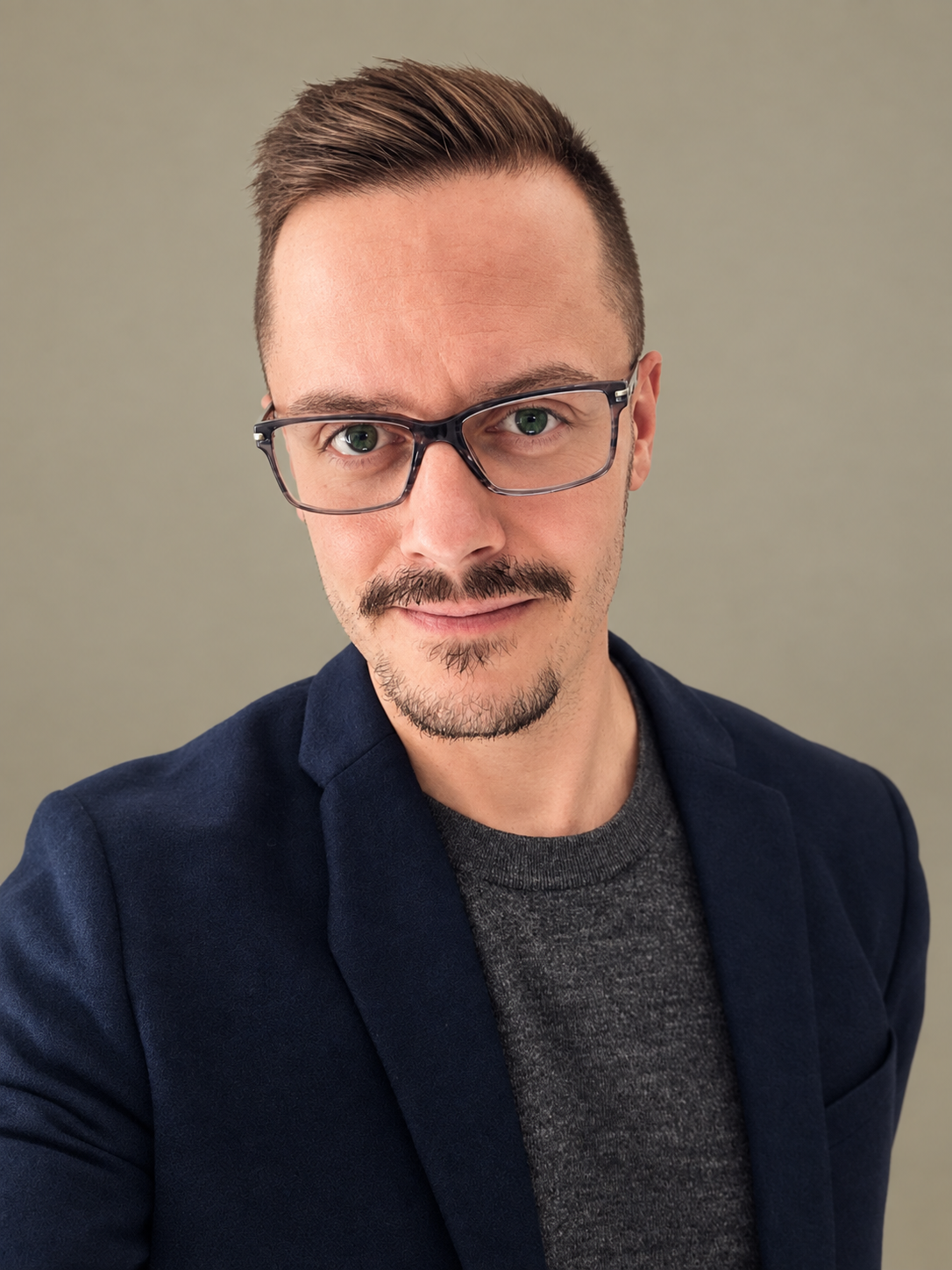 A close-up selfie of a man with short brown hair, green eyes, glasses, and a mustache, wearing a navy blazer and a gray shirt, against a plain light green background.