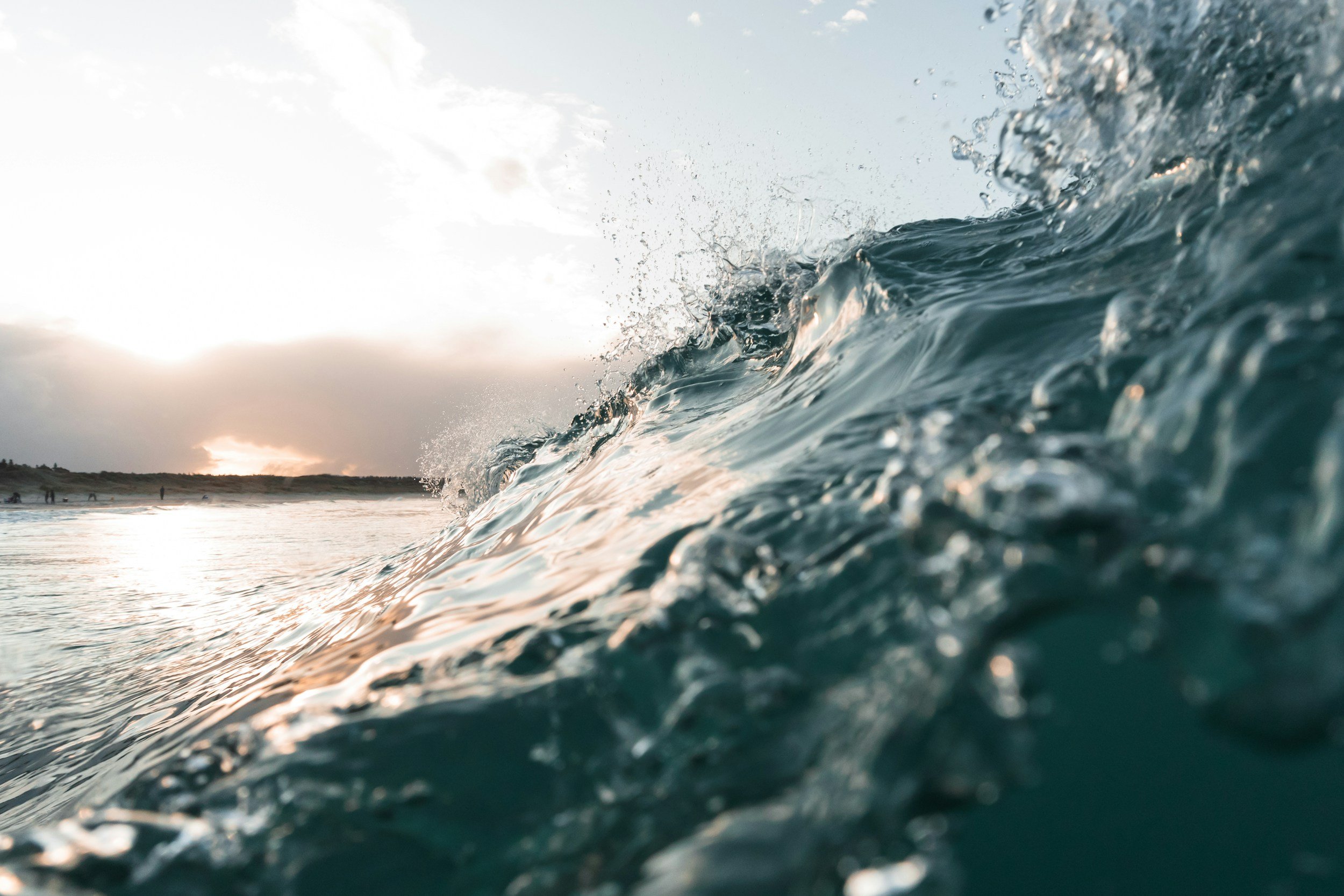 Close-up of ocean waves at sunset with people on the distant shoreline.