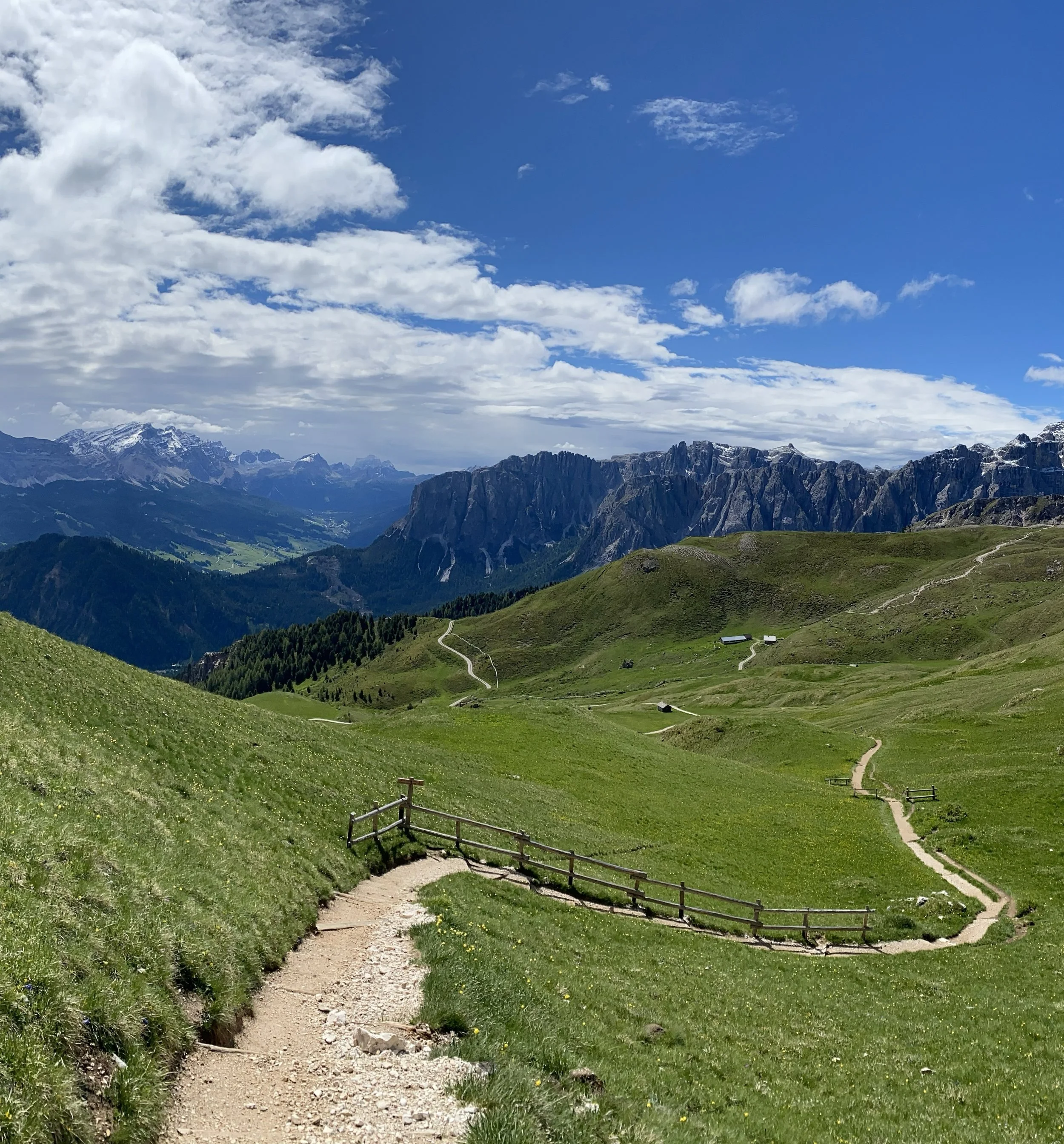 A scenic mountain landscape with green hills, winding pathways, and a distant mountain range with snow-capped peaks under a partly cloudy sky.