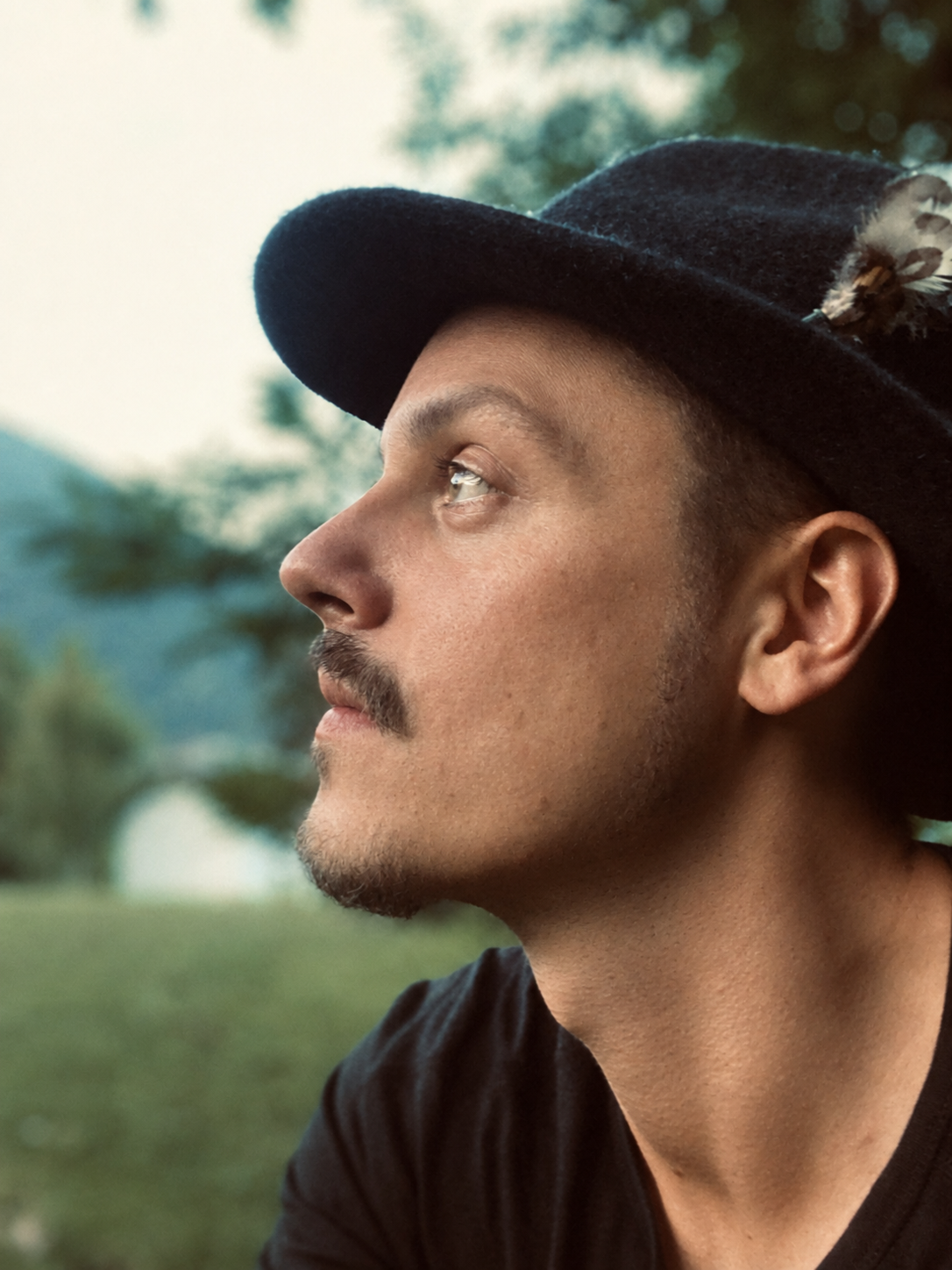 Side profile of a man wearing a black hat with feathers, looking thoughtfully into the distance outdoors with trees and mountains in the background.