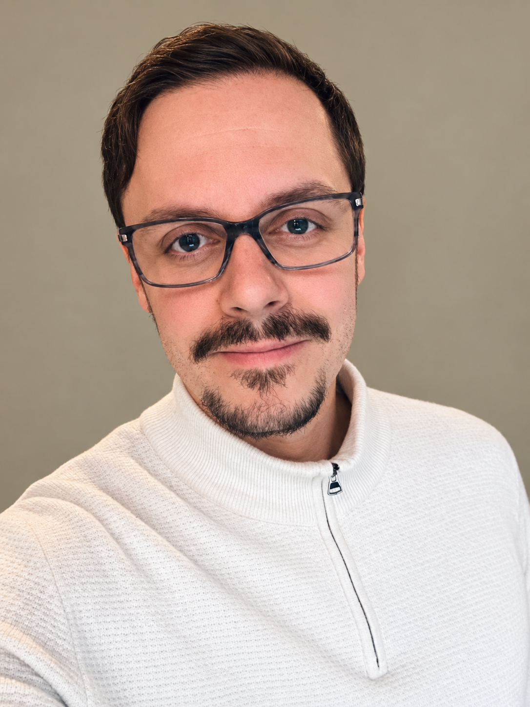 A man with brown hair, wearing glasses with dark frames, a mustache and goatee, in a white zip-up sweater, smiling at the camera.