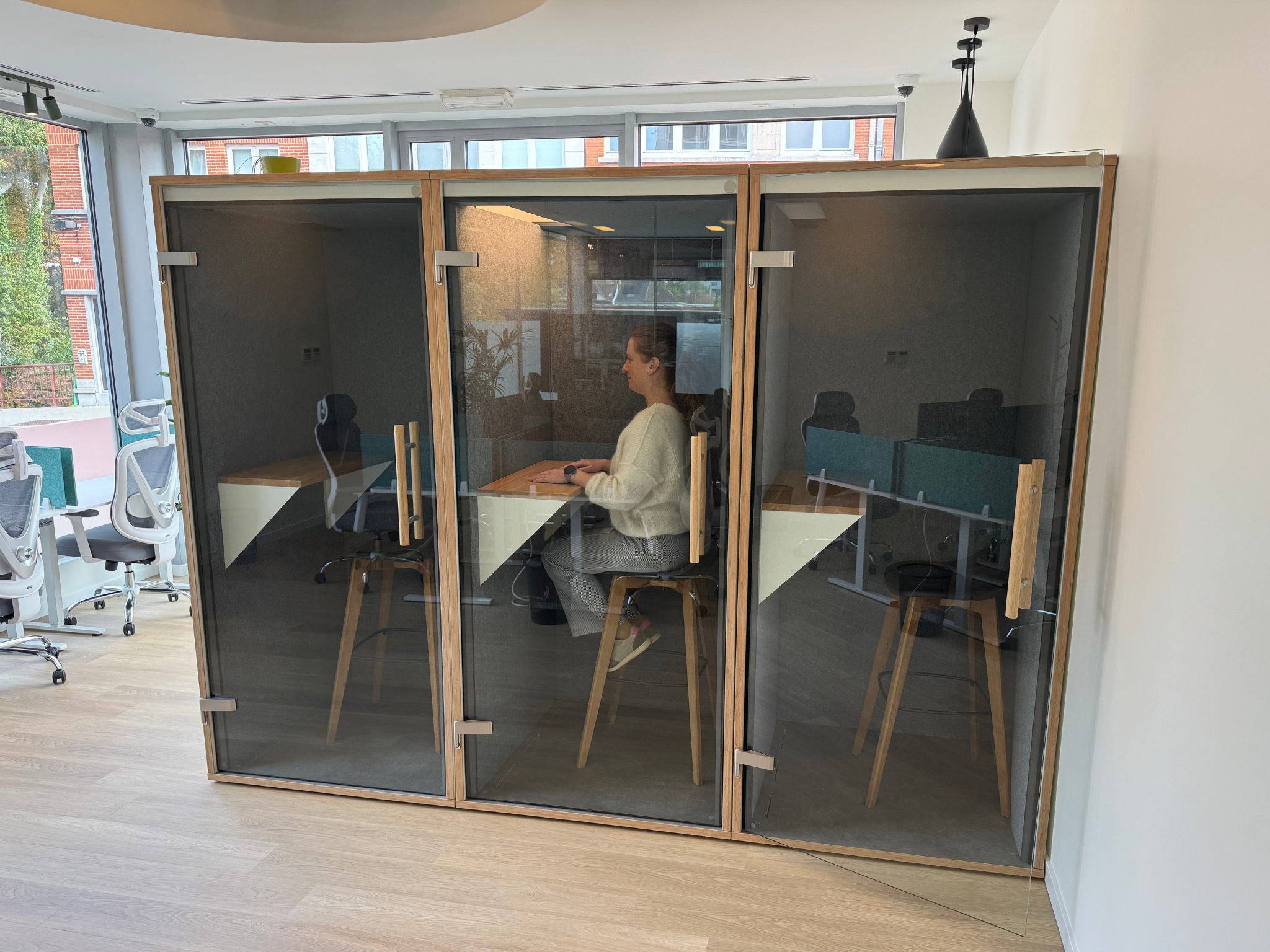 A woman sitting inside a soundproof phone booth in an office, with large windows, chairs, and office equipment visible outside.