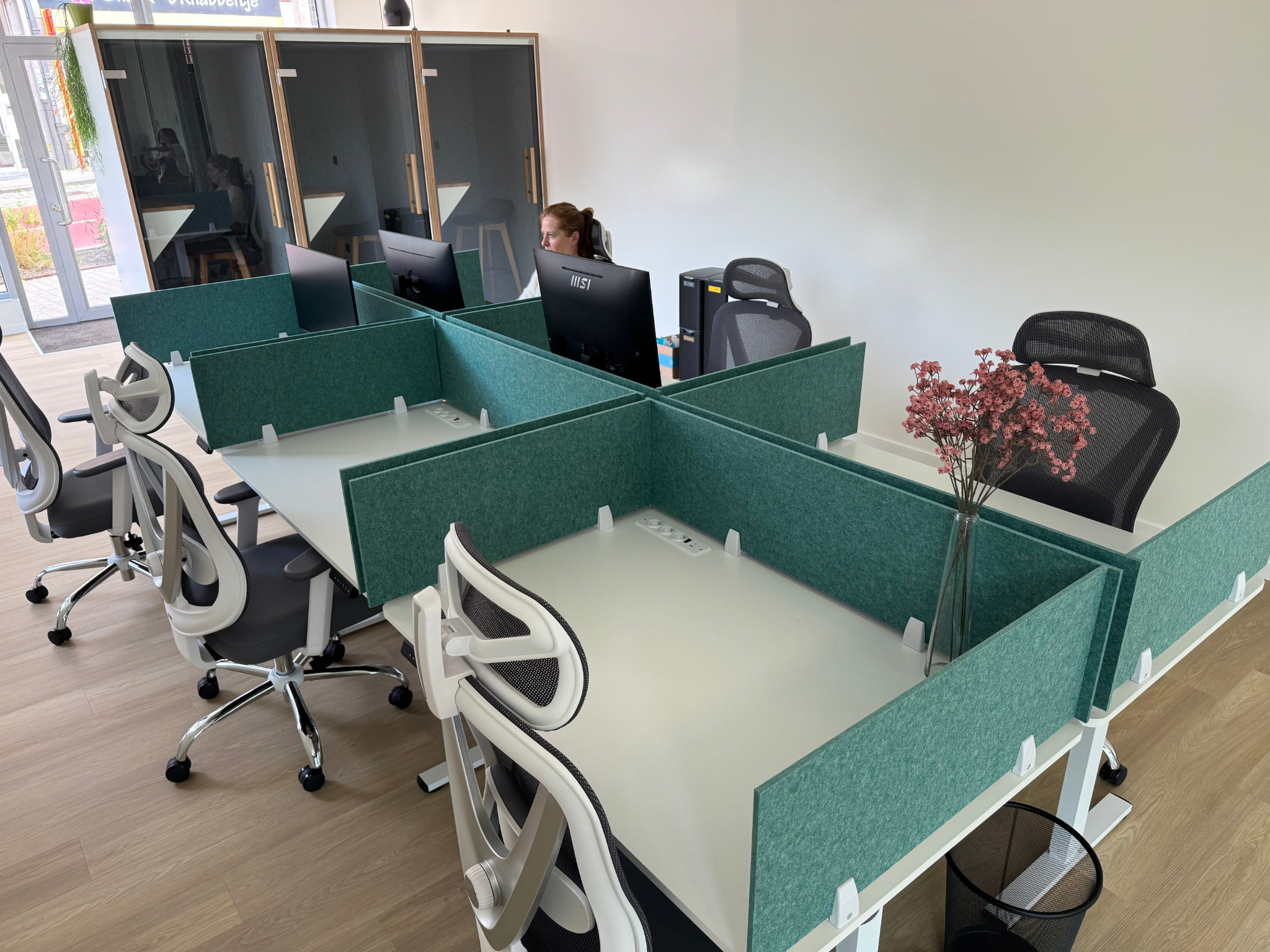 An office space with workstations separated by green fabric partitions. There are several ergonomic office chairs and computers on the desks. In the background, there are glass-walled private booths, and a woman is sitting at one of the computers. A 