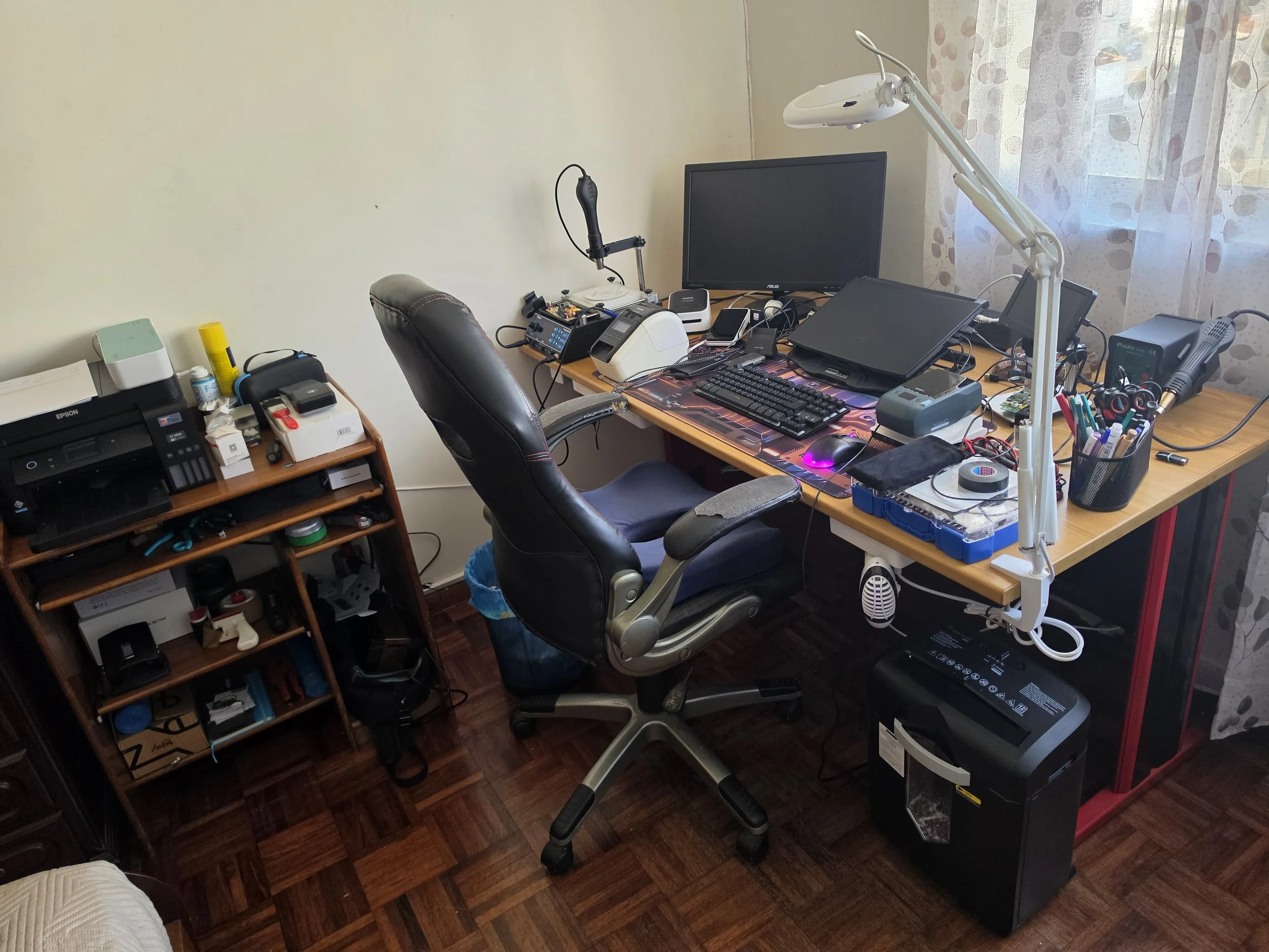 Escritório com mesa de madeira, cadeira de escritório, computador, microfones, monitor, impressora, luminária branca, vários cabos e materiais de escritório.