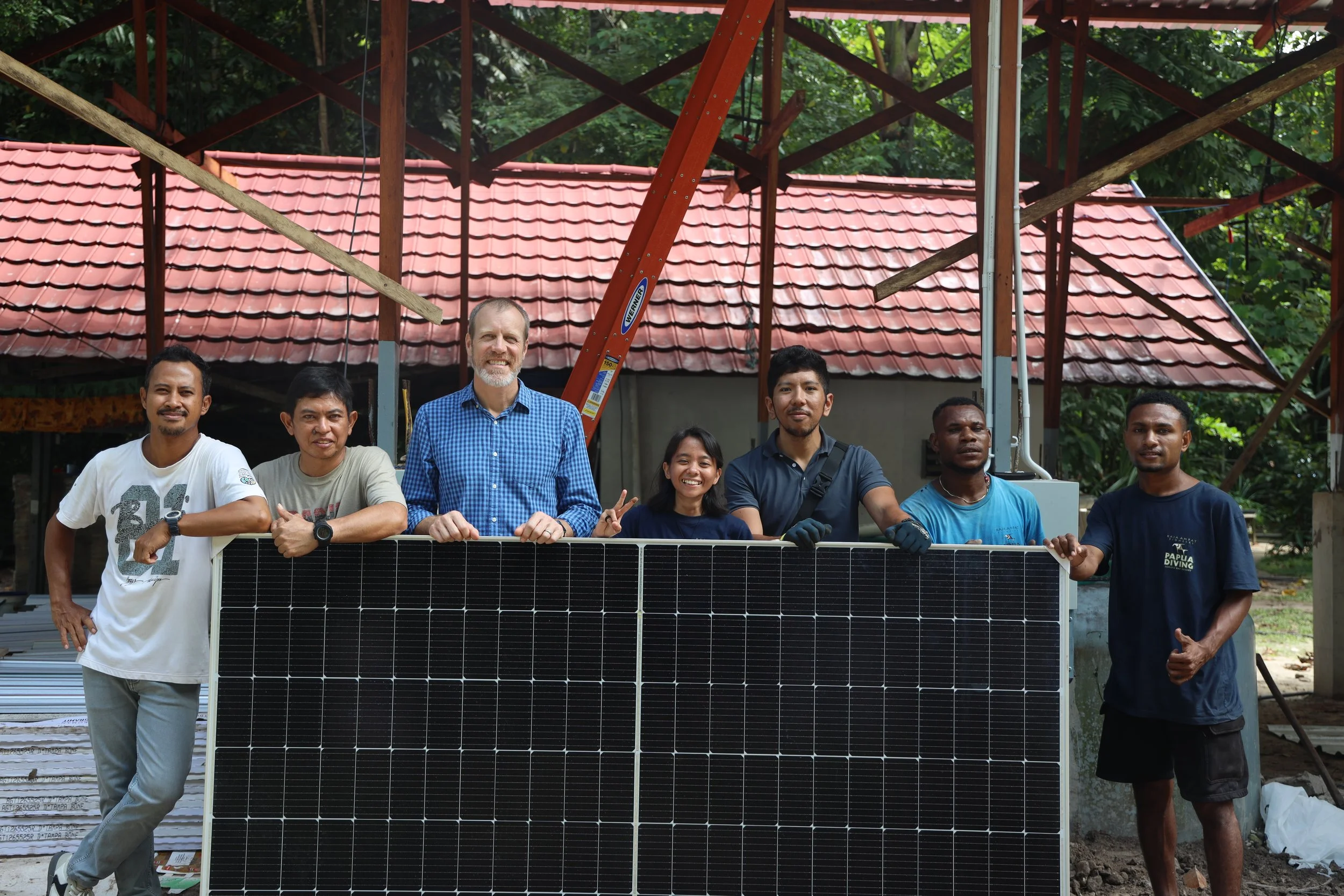 Group of seven diverse people standing outdoors behind a solar panel, with a construction site and trees in the background, some smiling and making peace signs.