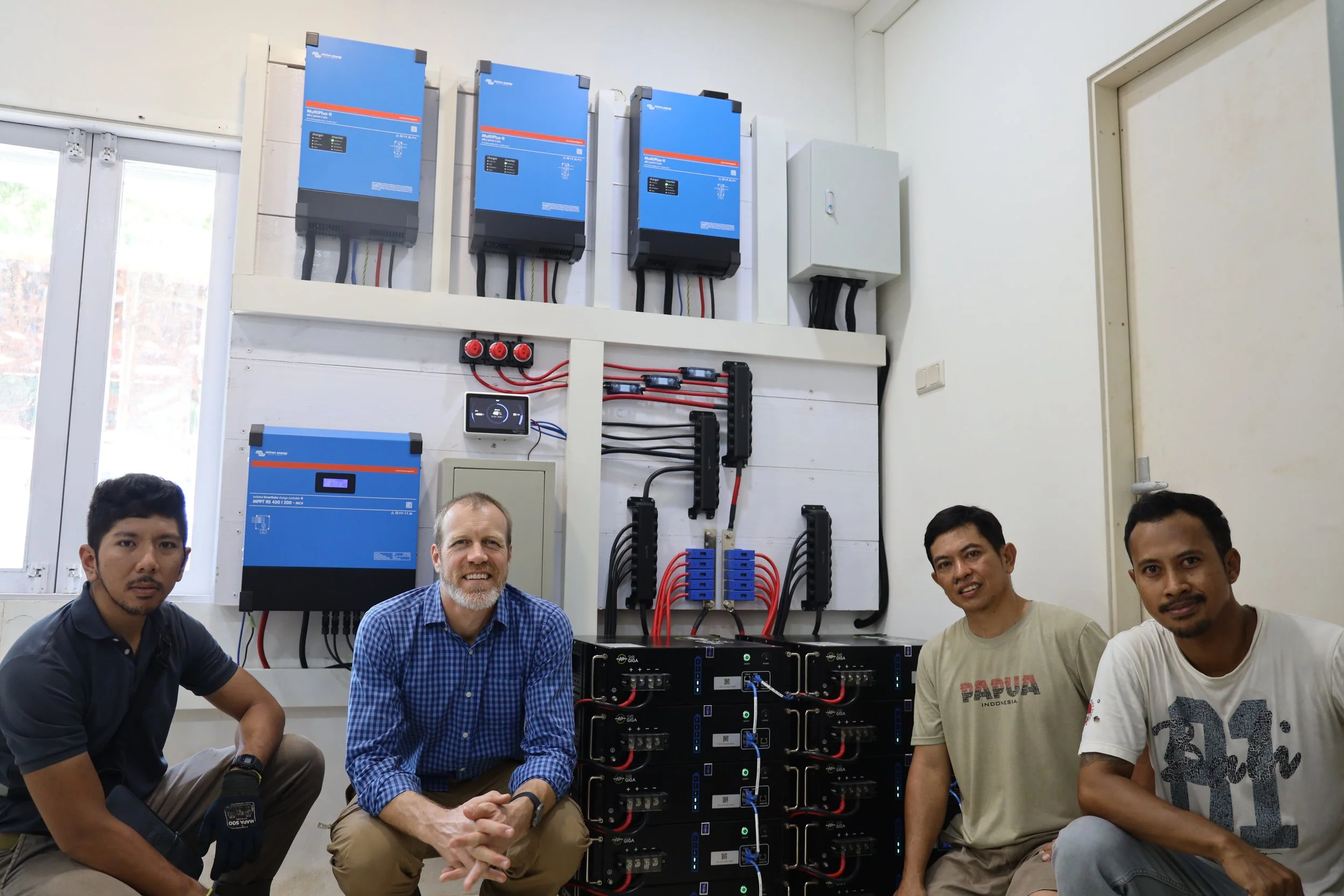 A group of five men, some smiling, are in a room with electronic equipment. They are sitting and kneeling in front of blue industrial devices mounted on the wall and connected with black and red cables. The room has a window on the left and a white wall on the right.