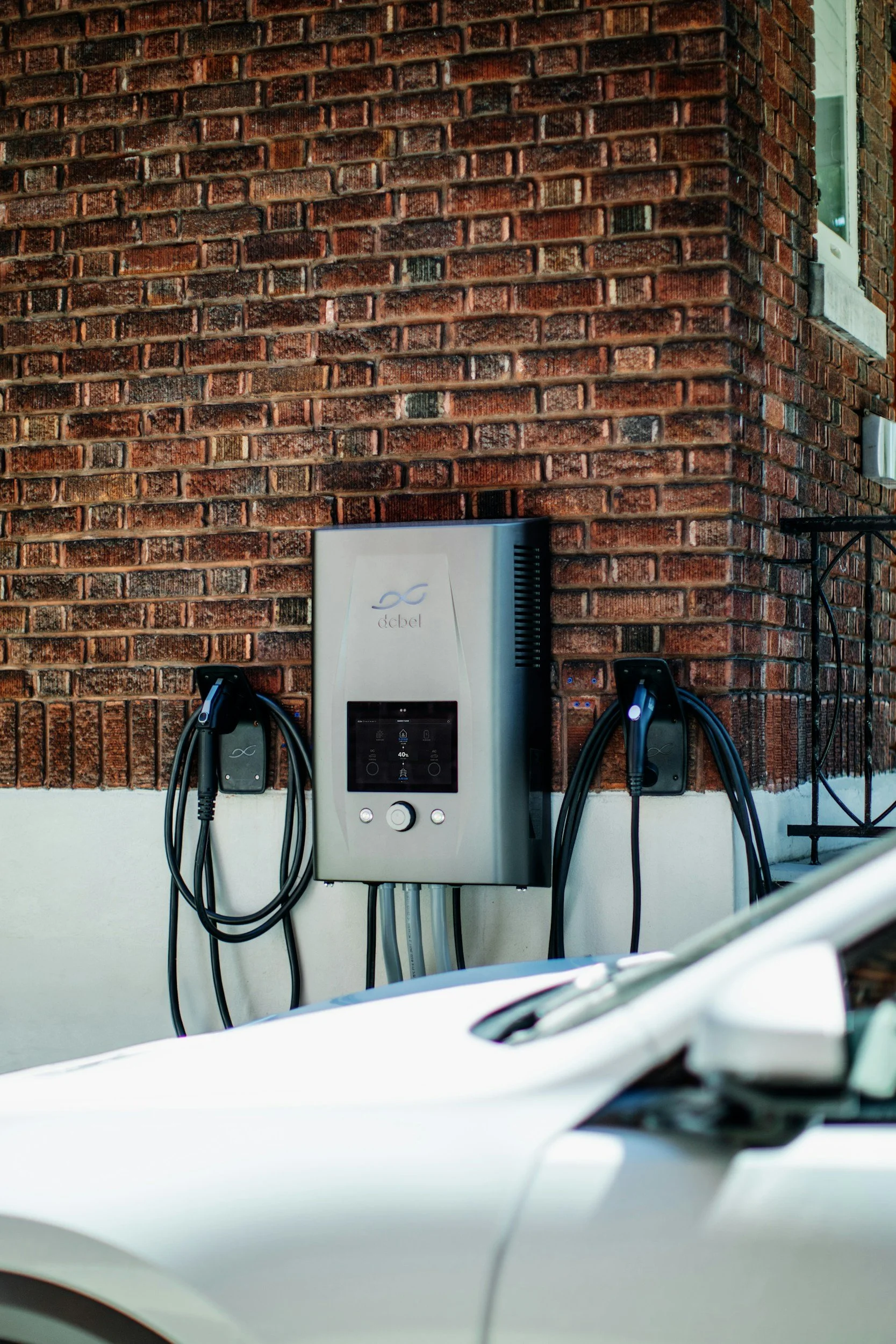Electric vehicle charging station mounted on a brick wall, with black charging cables hanging from it, and a white vehicle partially visible in the foreground.