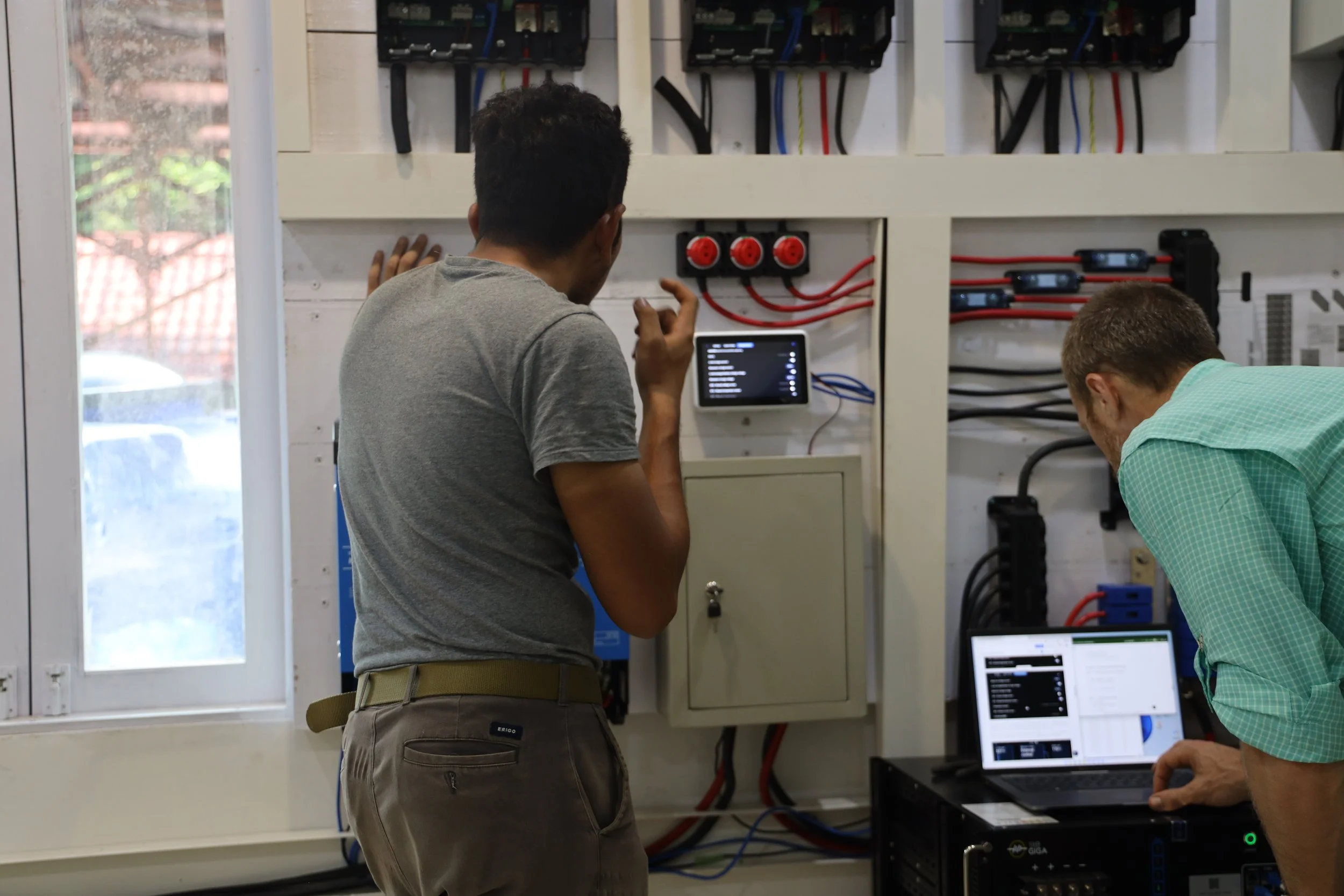 Two men working on electrical panels with wires and a laptop in a room with a window.