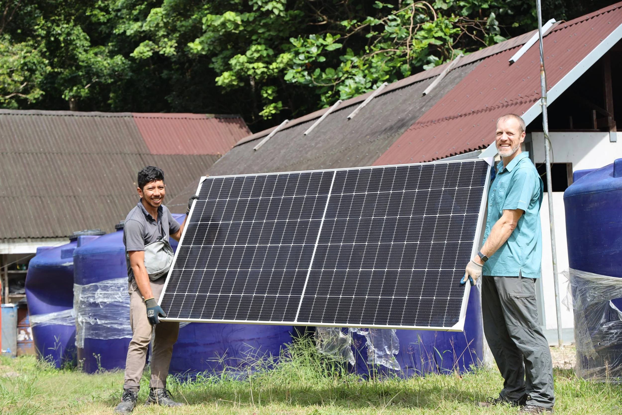 Two men are posing outdoors with a solar panel, with one man holding the panel. They are standing in front of a rustic building with a red and gray roof and blue water tanks. The background features lush green trees.