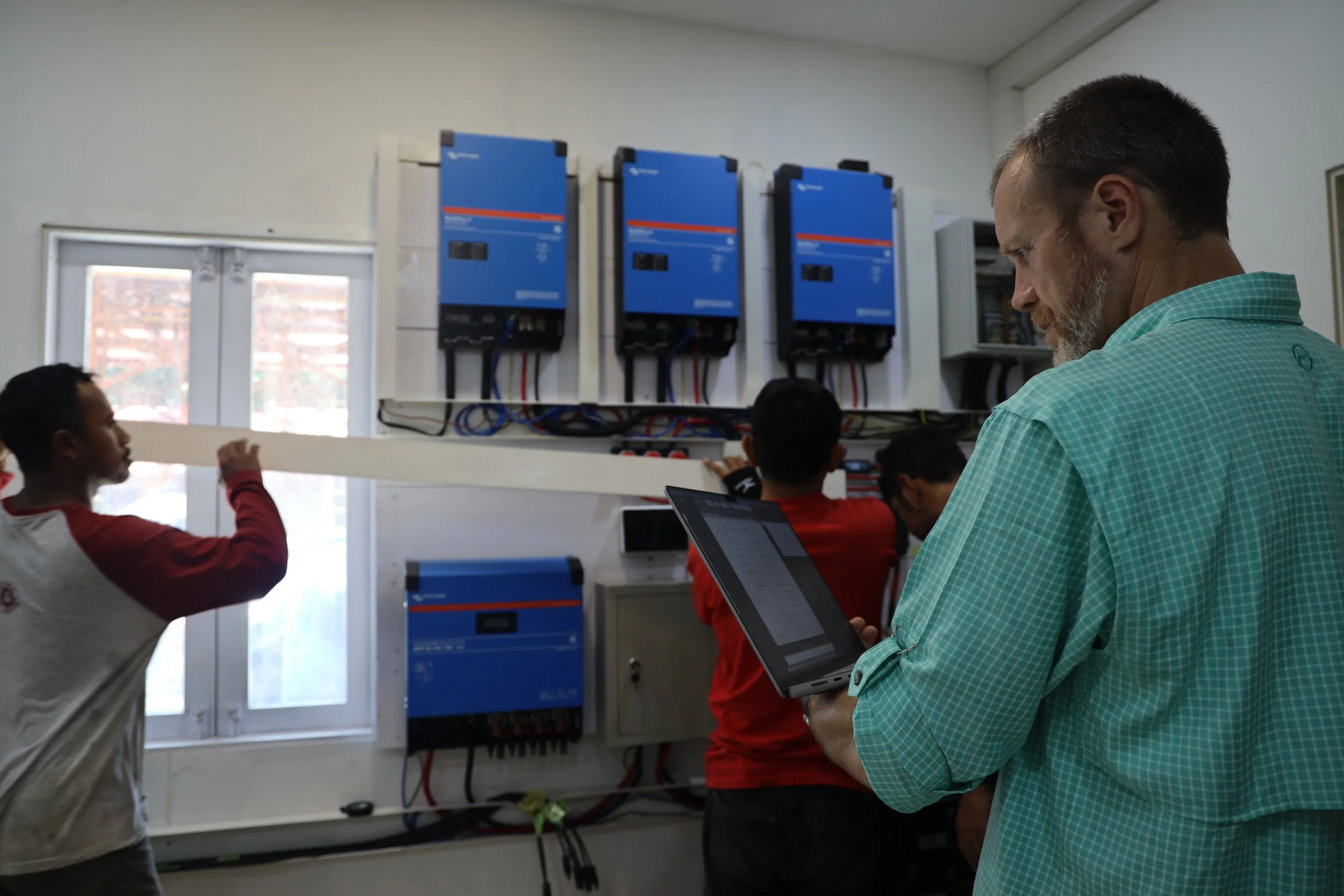 A man with a beard and glasses holding a tablet in a room with blue electrical equipment on the wall and children sitting at a table.