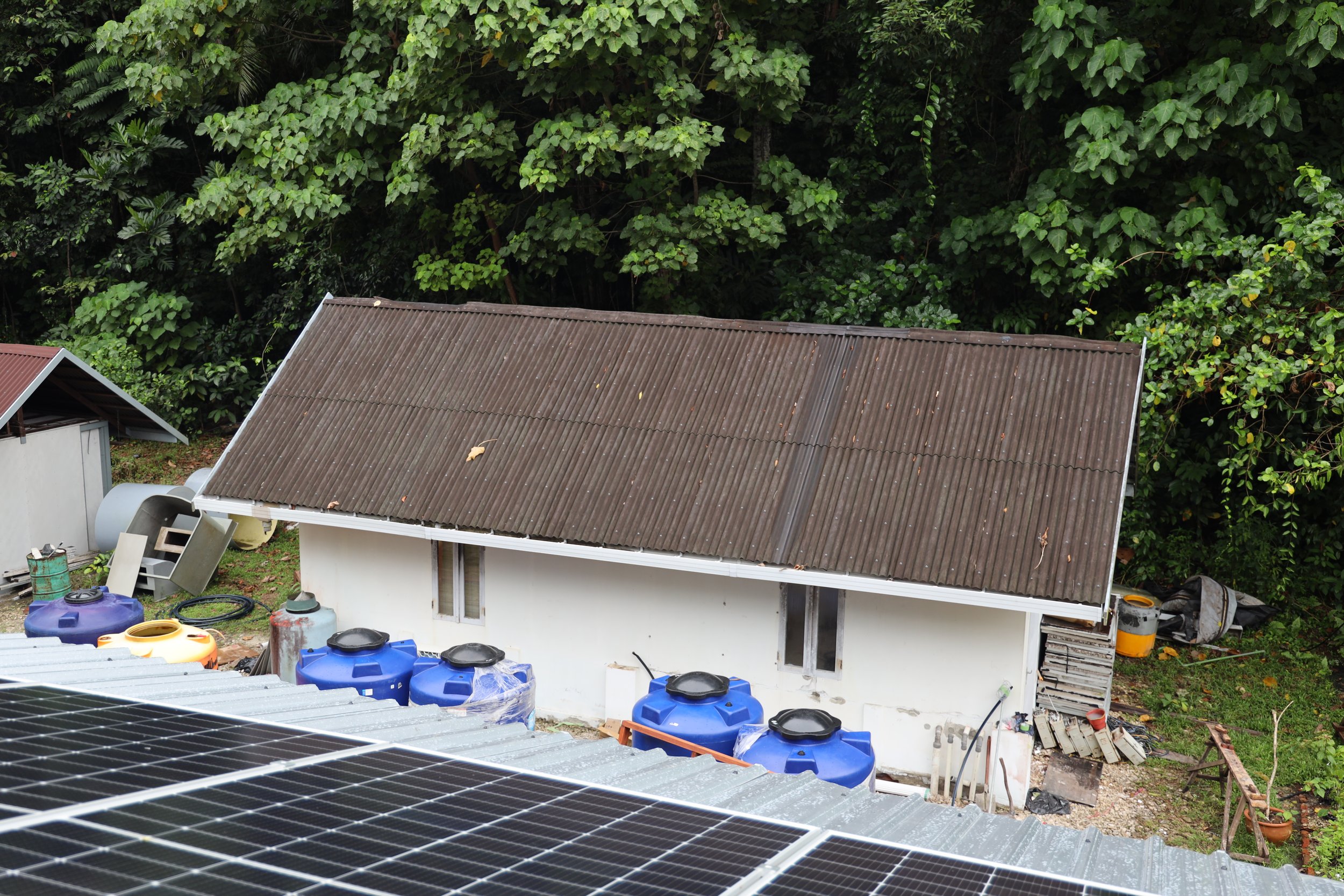 View of a backyard with a house featuring a sloped, ribbed roof, surrounded by dense green foliage and trees in the background. The yard has various containers and objects, including blue water tanks and solar panels in the foreground.