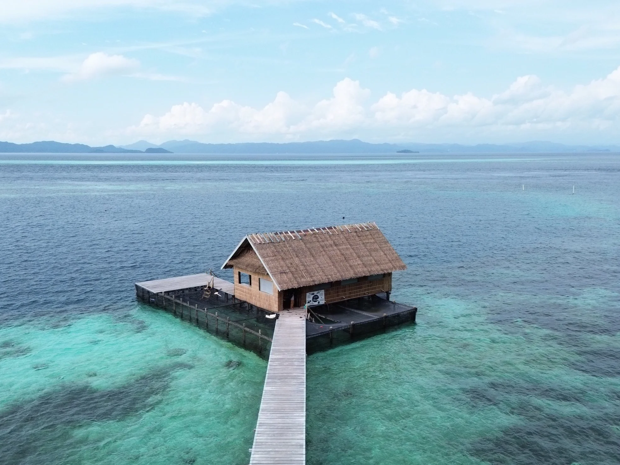 A thatched-roof hut built on stilts over a clear, turquoise ocean with a pier leading to the hut and mountains in the distant background.
