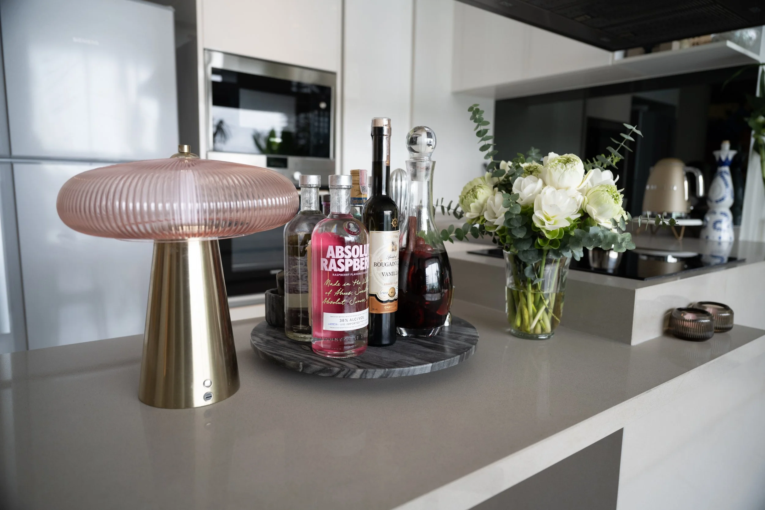 Grey marble 'lazy-susan' tray of spirits and alcohol drinks, beside a pink and gold ribbed-glass mushroom map and a bouquet of white lotus flowers