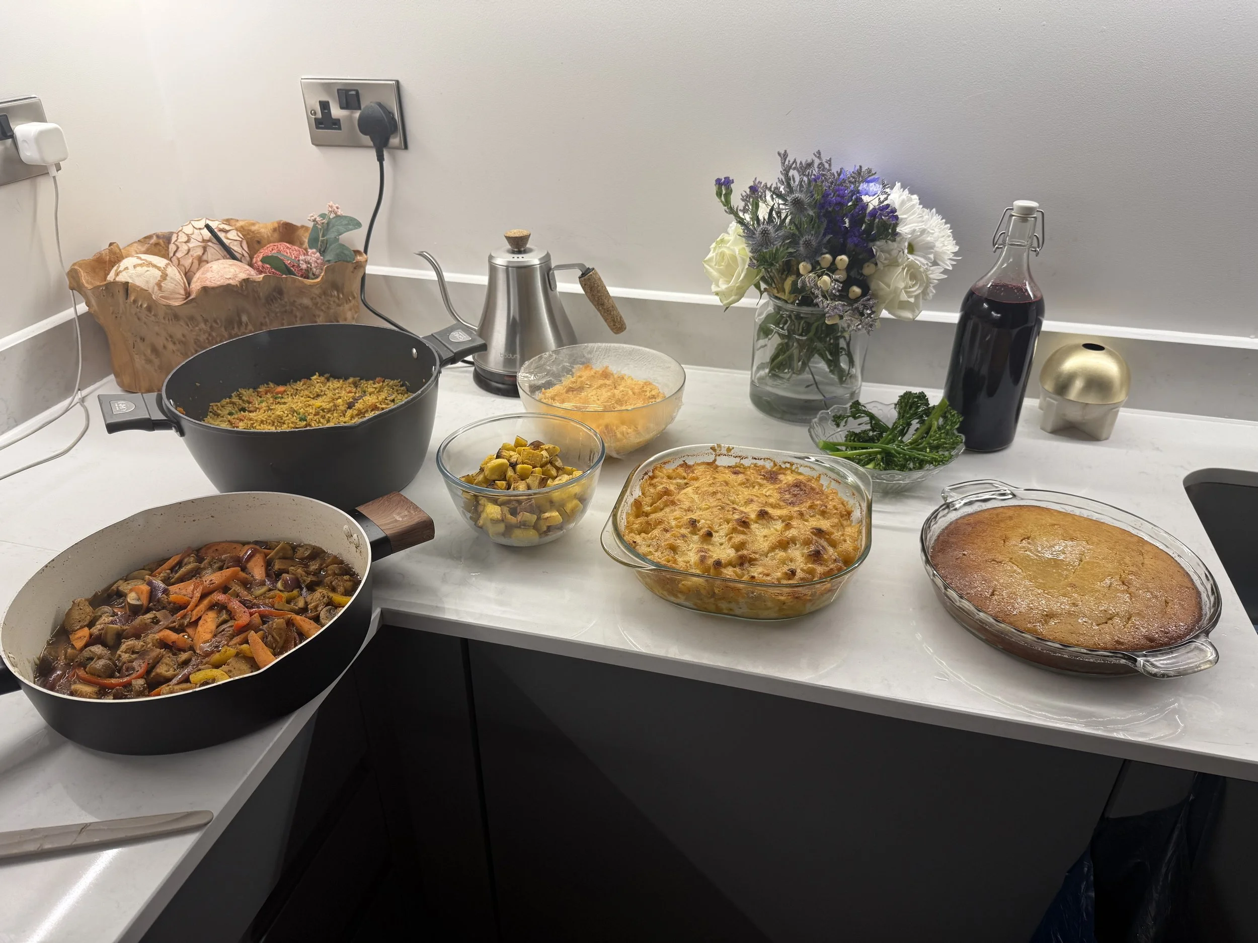 Kitchen countertop with pots of delicious vegan food including fried rice, jerk vegetables, mac and cheese, fried plantain, homemade coleslaw, honey-butter cornbread and sautéed broccolini