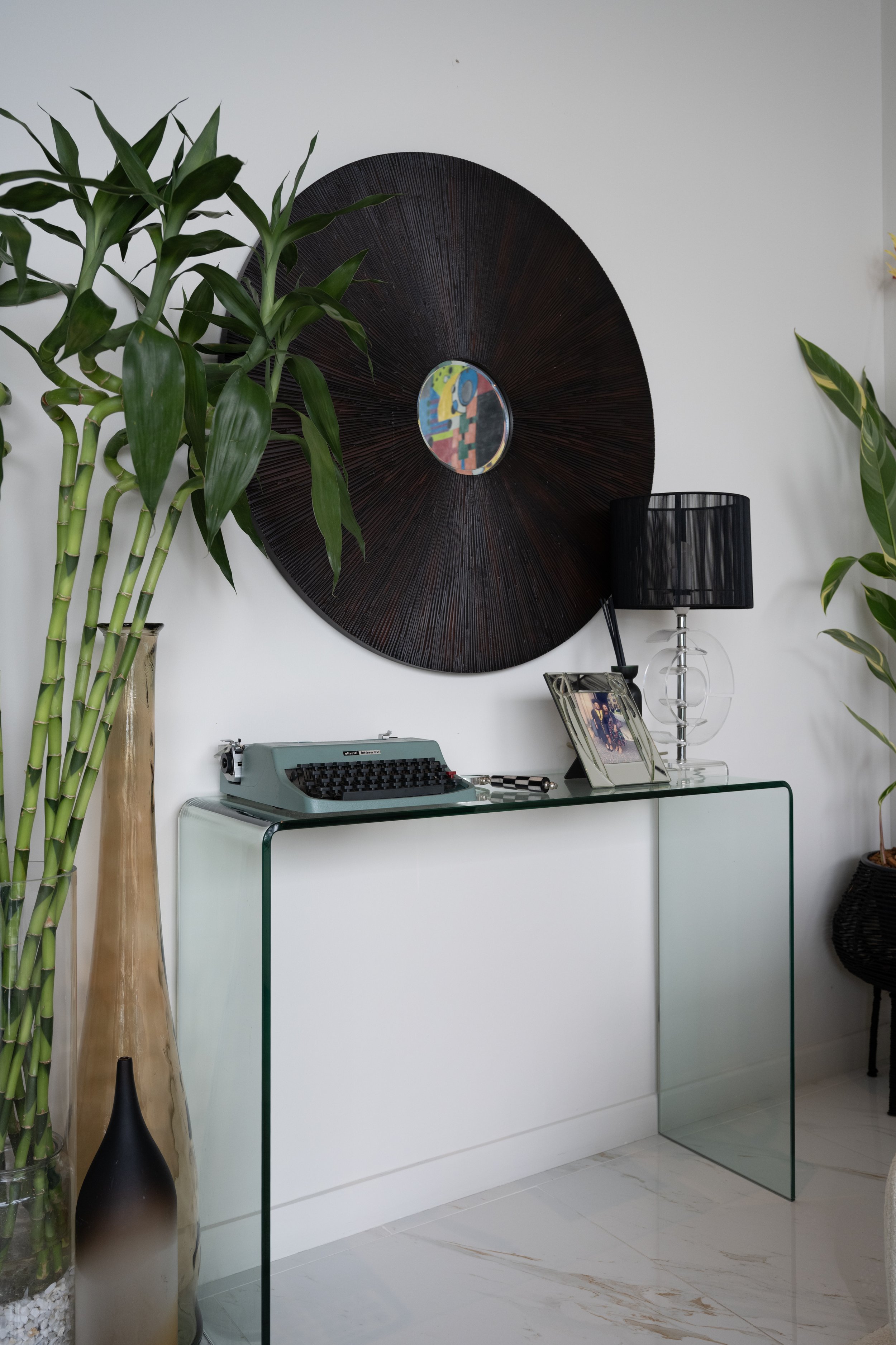 Vintage blue typewriter on a clear glass console table beside tall lucky bamboos and a large round wooden wall mirror 