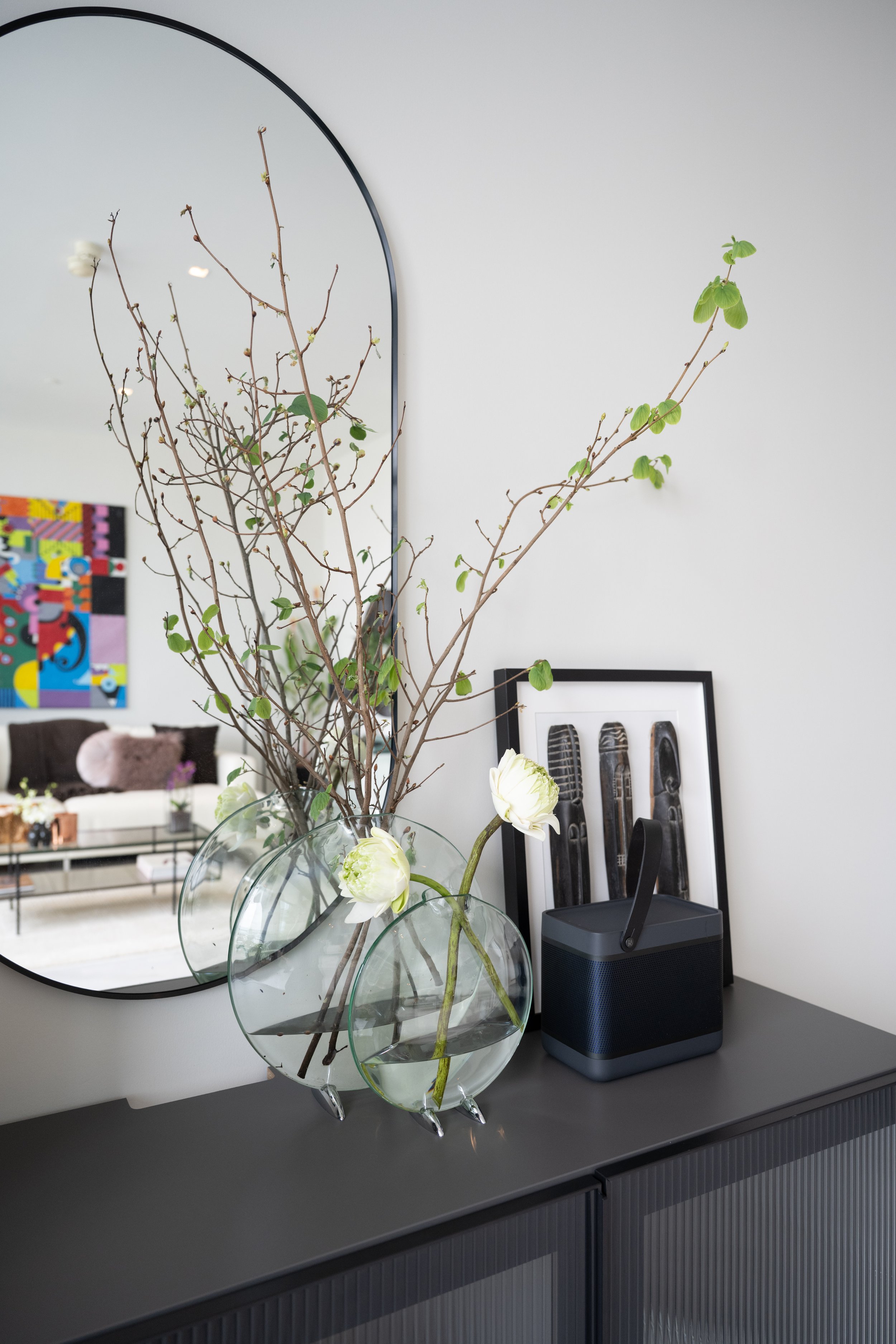 Budding branches and white lotus flowers in round glass vases before an arched mirror reflecting a colourful painting