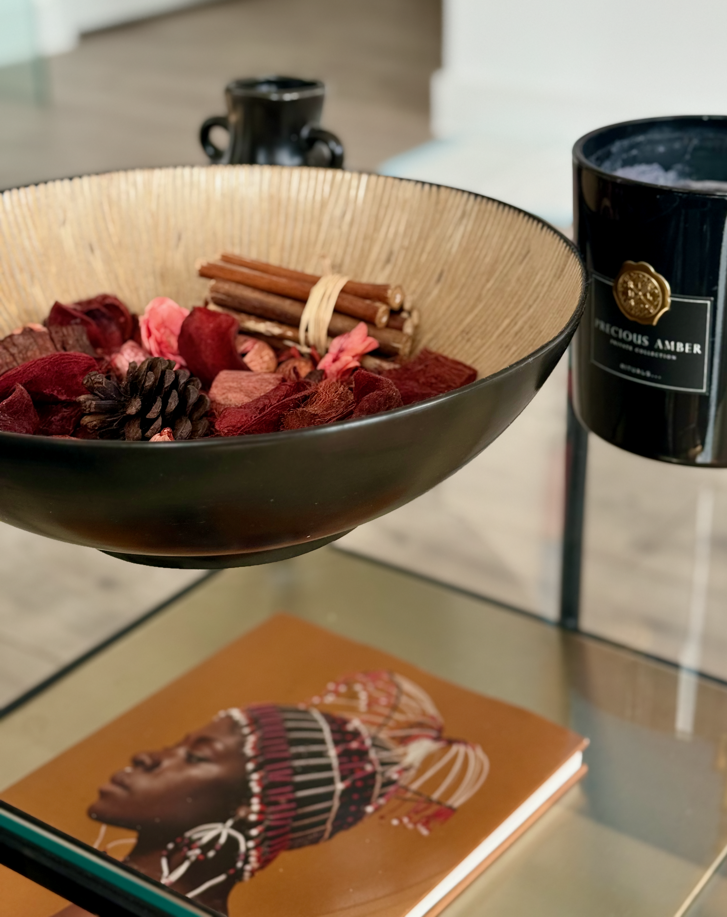 Bowl of pink and red pot-pourri with cinnamon sticks, beside an extra large black candle and a coffee table book with a Black woman with an elaborate beaded headpiece on the cover