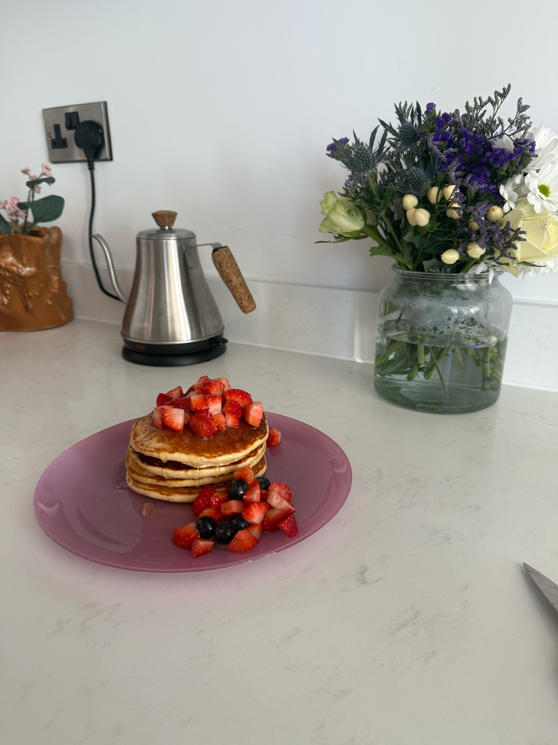 Fluffy pancakes served with slices of fresh strawberries, blueberries and honey. Plated on a glass pink plate with a purple bouquet behind
