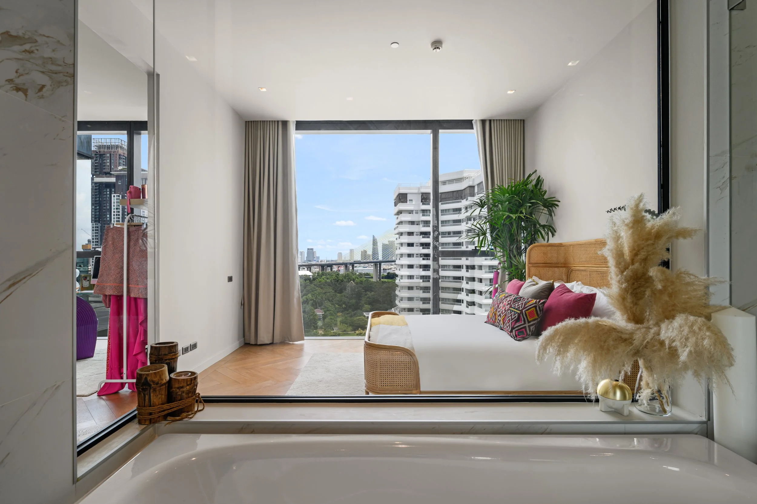 Rattan bed with white bedding, pink velvet cushions facing floor-to-ceiling windows and a Bangkok skyline, seen from the bath