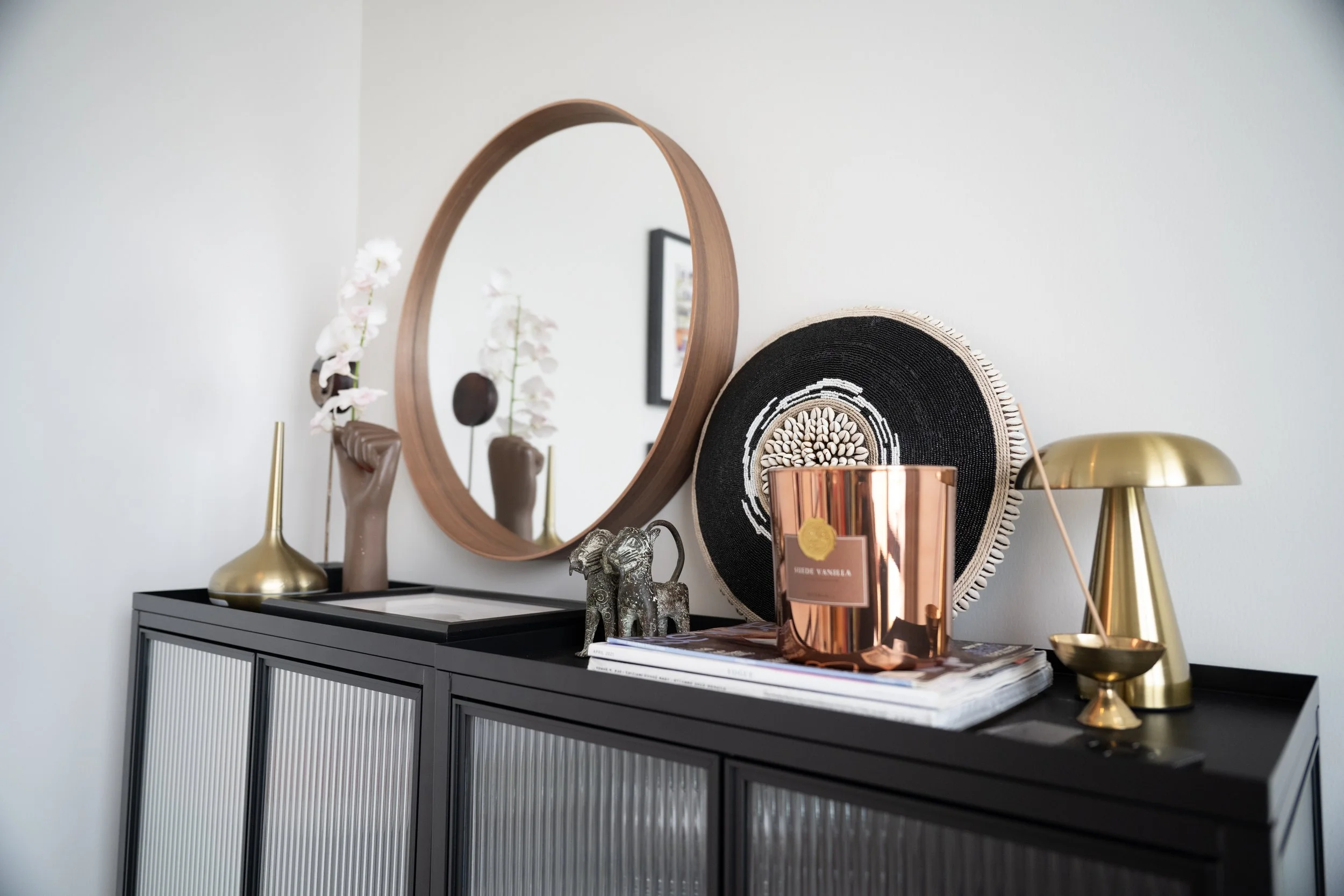 Beaded cowrie shell shield, bronze leopard figures, a raised fist vase holding white orchids and a copper candle on a black sideboard