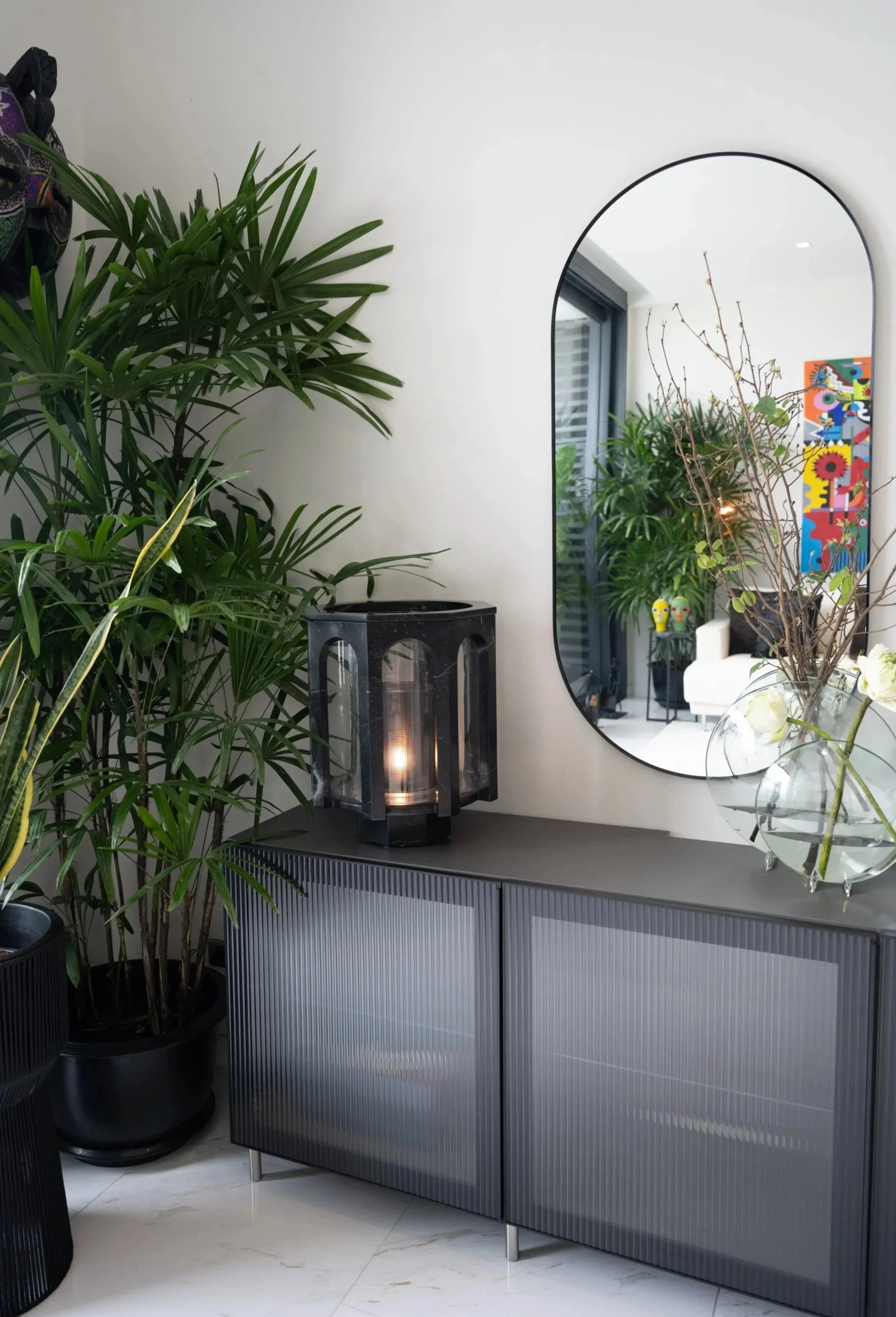 Black marble lantern lamp with arched panels, on a fluted dark grey sideboard beside a tall palm