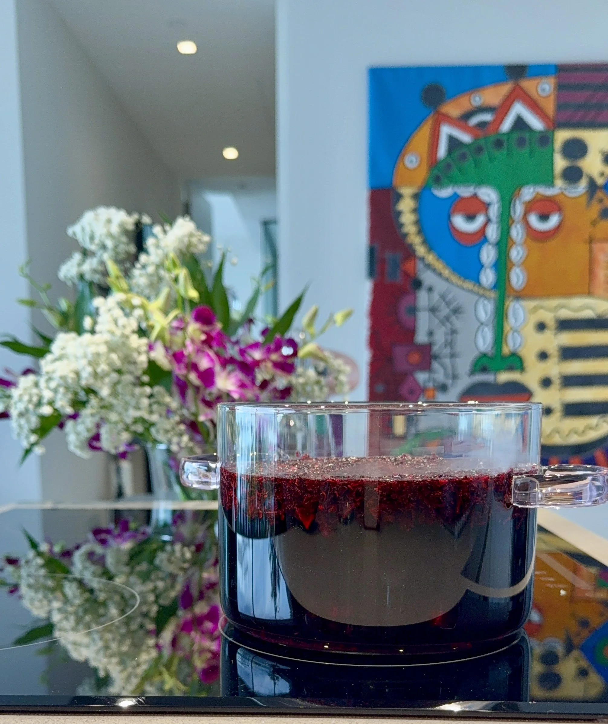 Dried hibiscus petals steeping in a clear pot. Behind is a purple and white bouquet of flowers and colourful wall art.