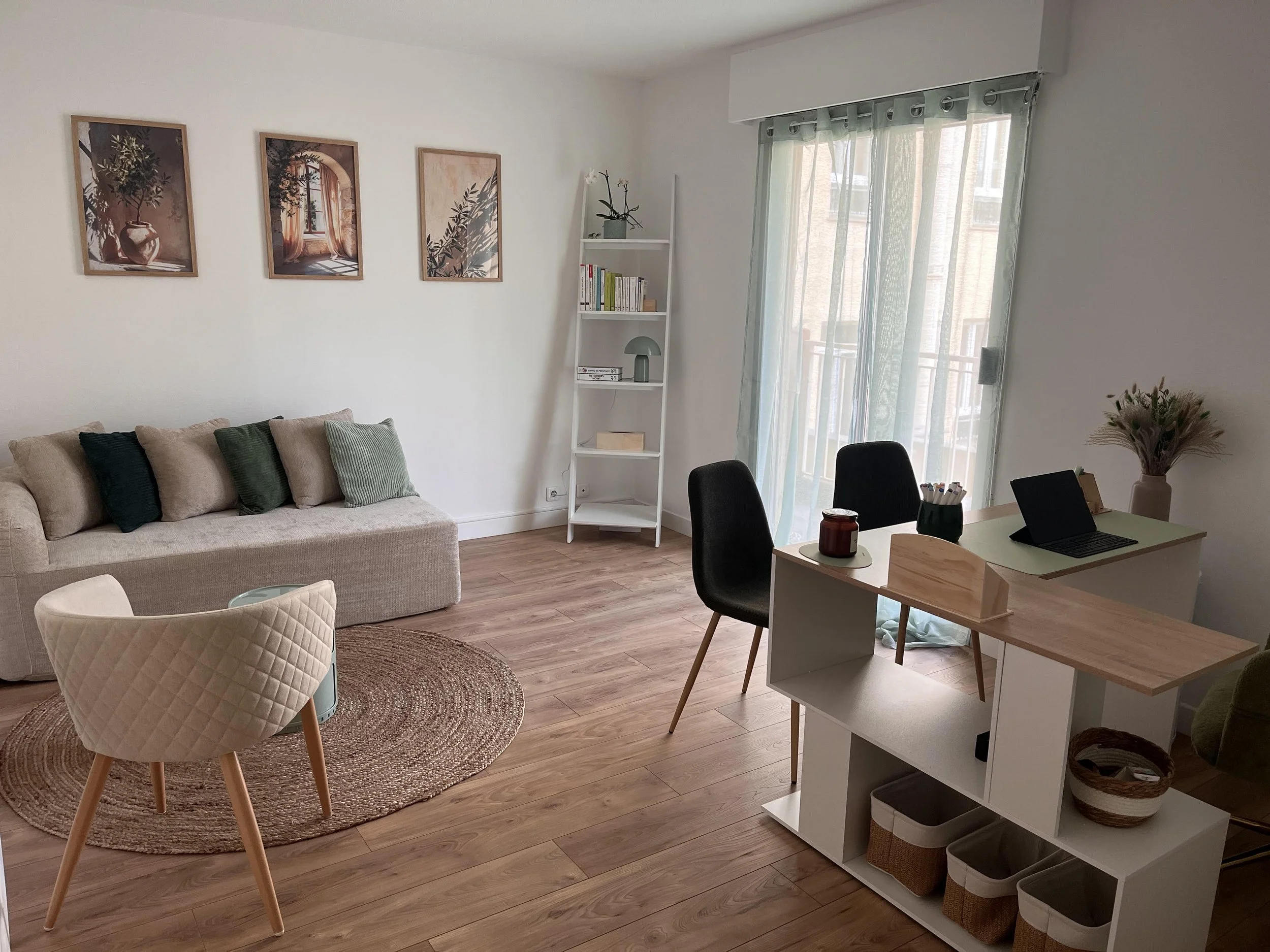 Salon avec un canapé beige, une table d'appoint, une étagère blanche, une table avec un ordinateur portable, des chaises, une fenêtre avec des voilages, des tableaux sur le mur, et un tapis rond en rotin.