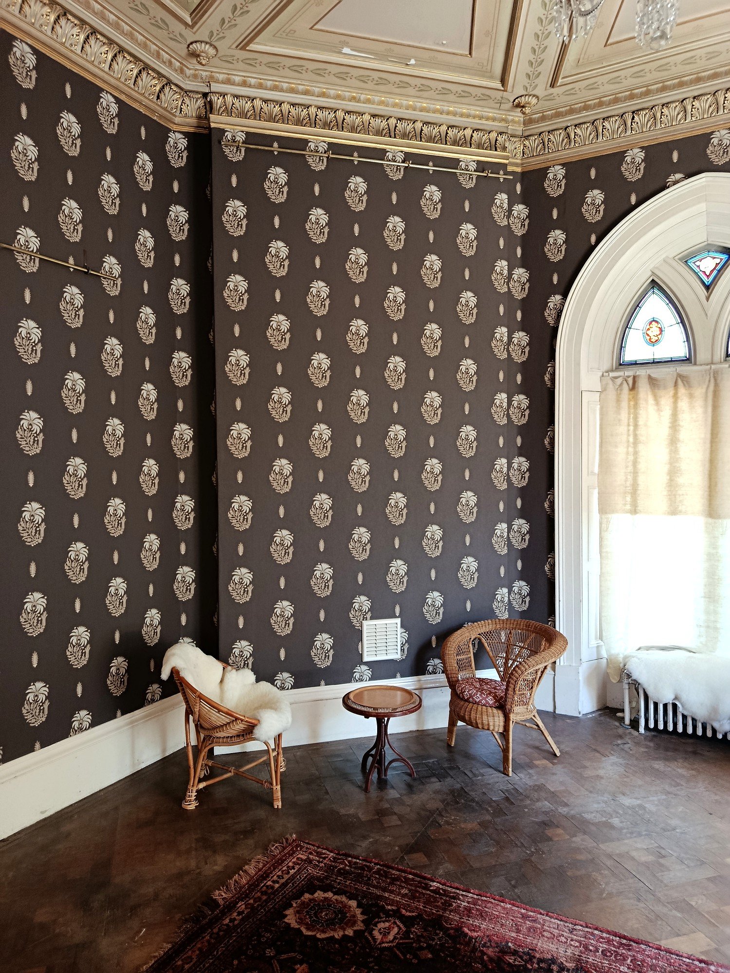 Dark wallpaper with cream motifs fitted around a room corner beside an arched window, beneath ornate cornicing.
