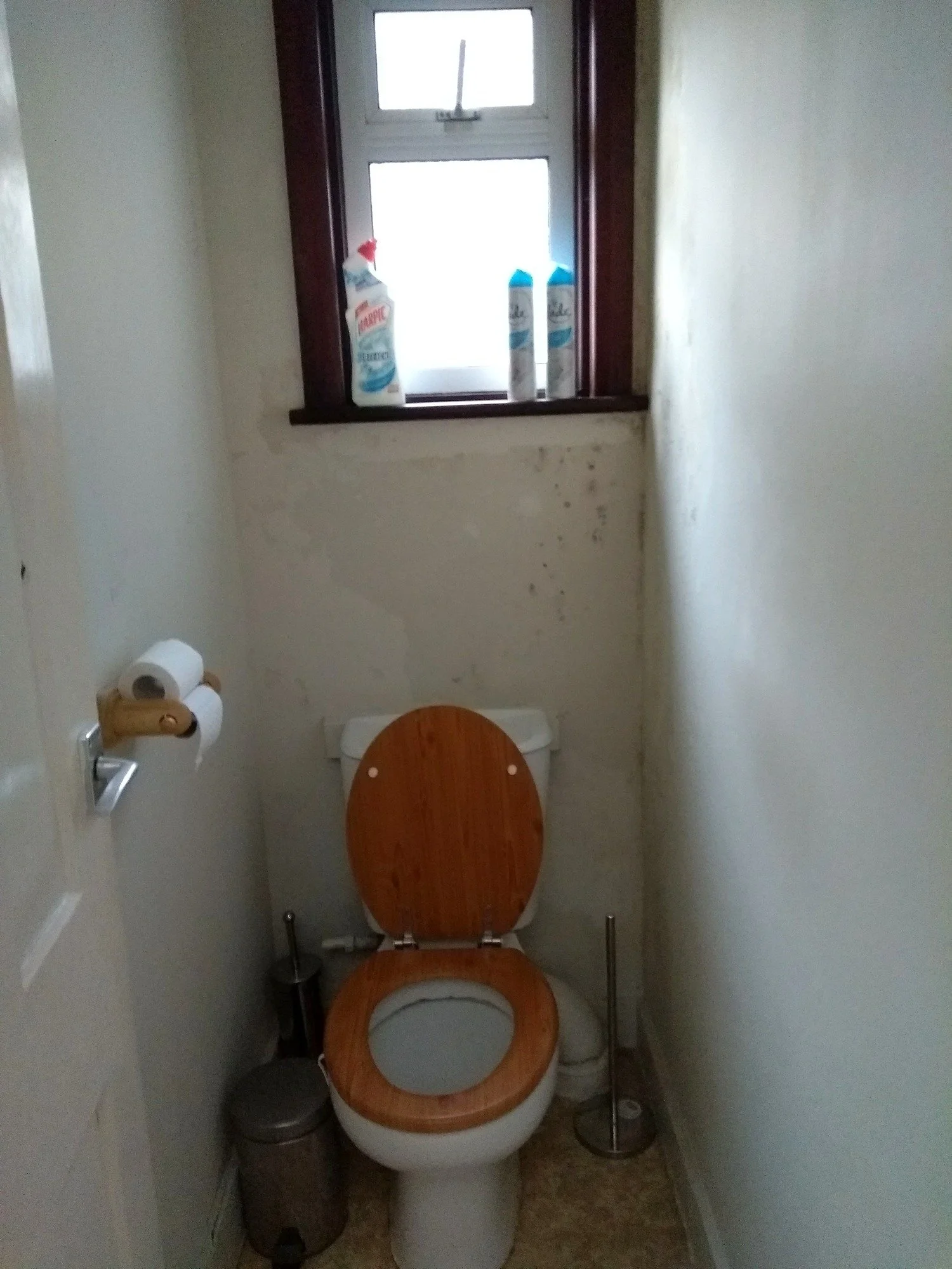 Separate toilet before refurbishment, with plain walls, a dark window frame and a wooden toilet seat.