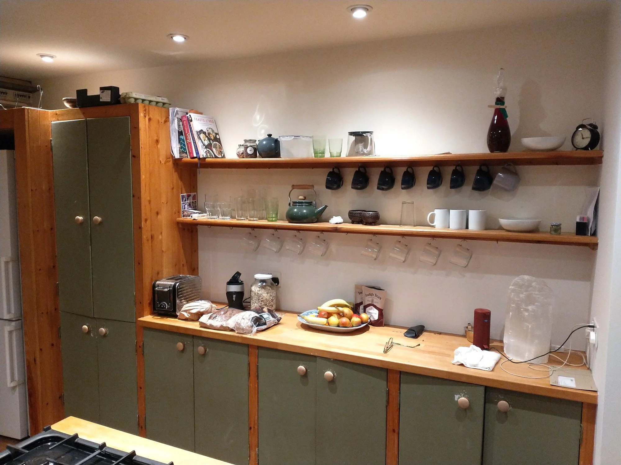 Kitchen before painting, with green cupboard doors, exposed timber cabinet frames and wooden wall shelves.