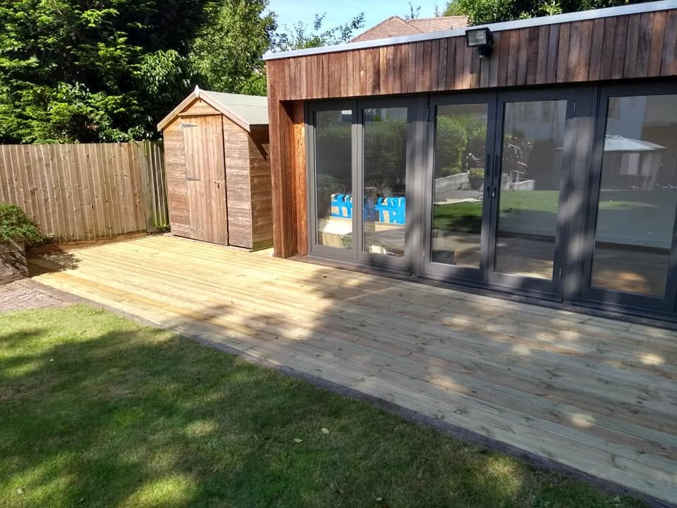 New timber decking extending across the front of a garden room and adjoining shed.