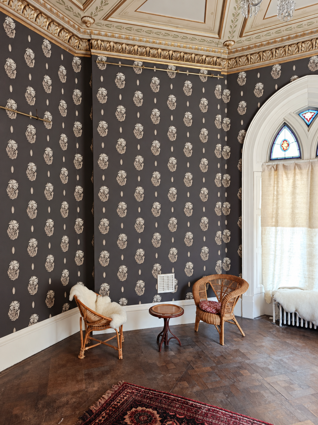 Room with dark patterned wallpaper, ornate cornicing and a white arched window with stained glass.