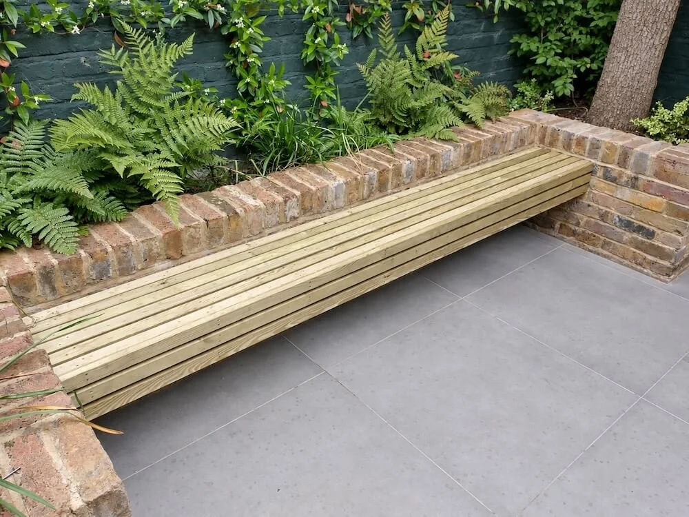 A wooden bench built along a brick flower bed in a garden with ferns and green plants, with gray tiled flooring.