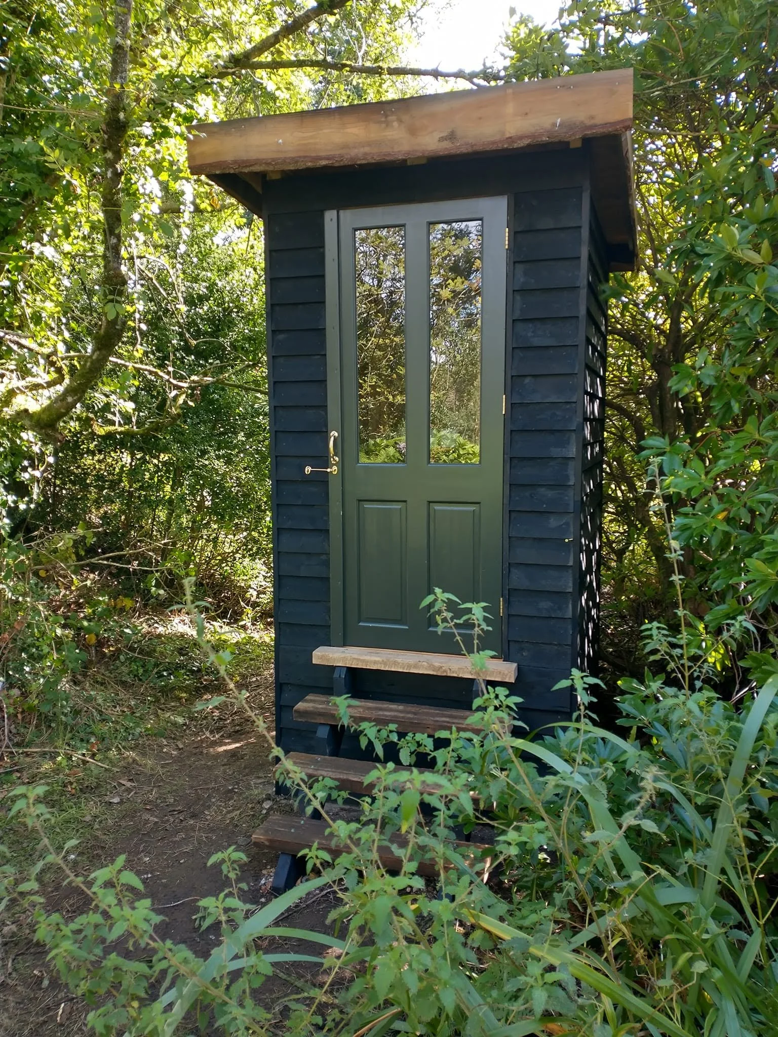 Dark-painted timber exterior of the garden composting toilet, with a glazed door beneath an overhanging roof.