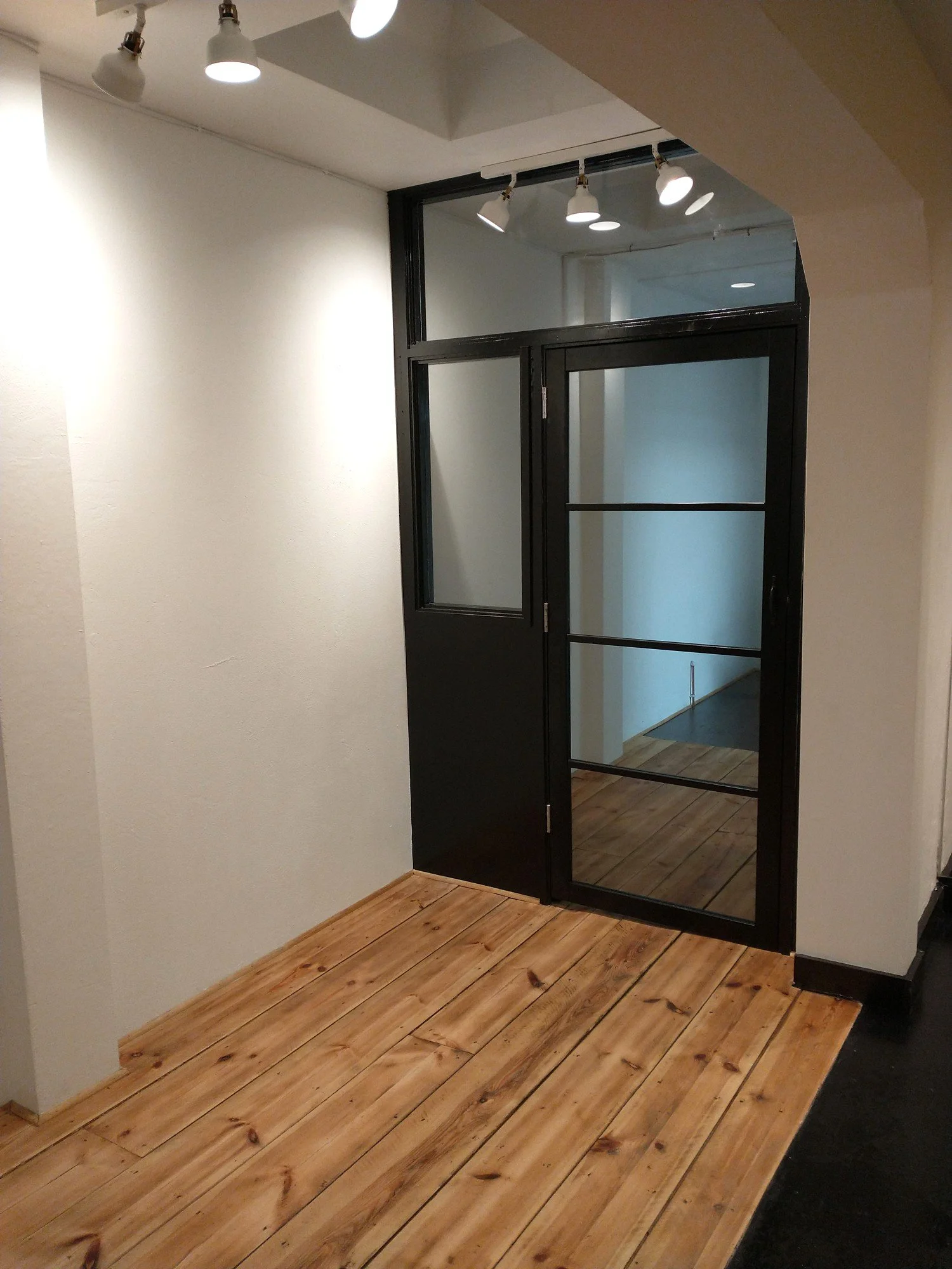 Black-framed glazed doorway with an overhead glass panel, white walls and timber floorboards in the refurbished shop.