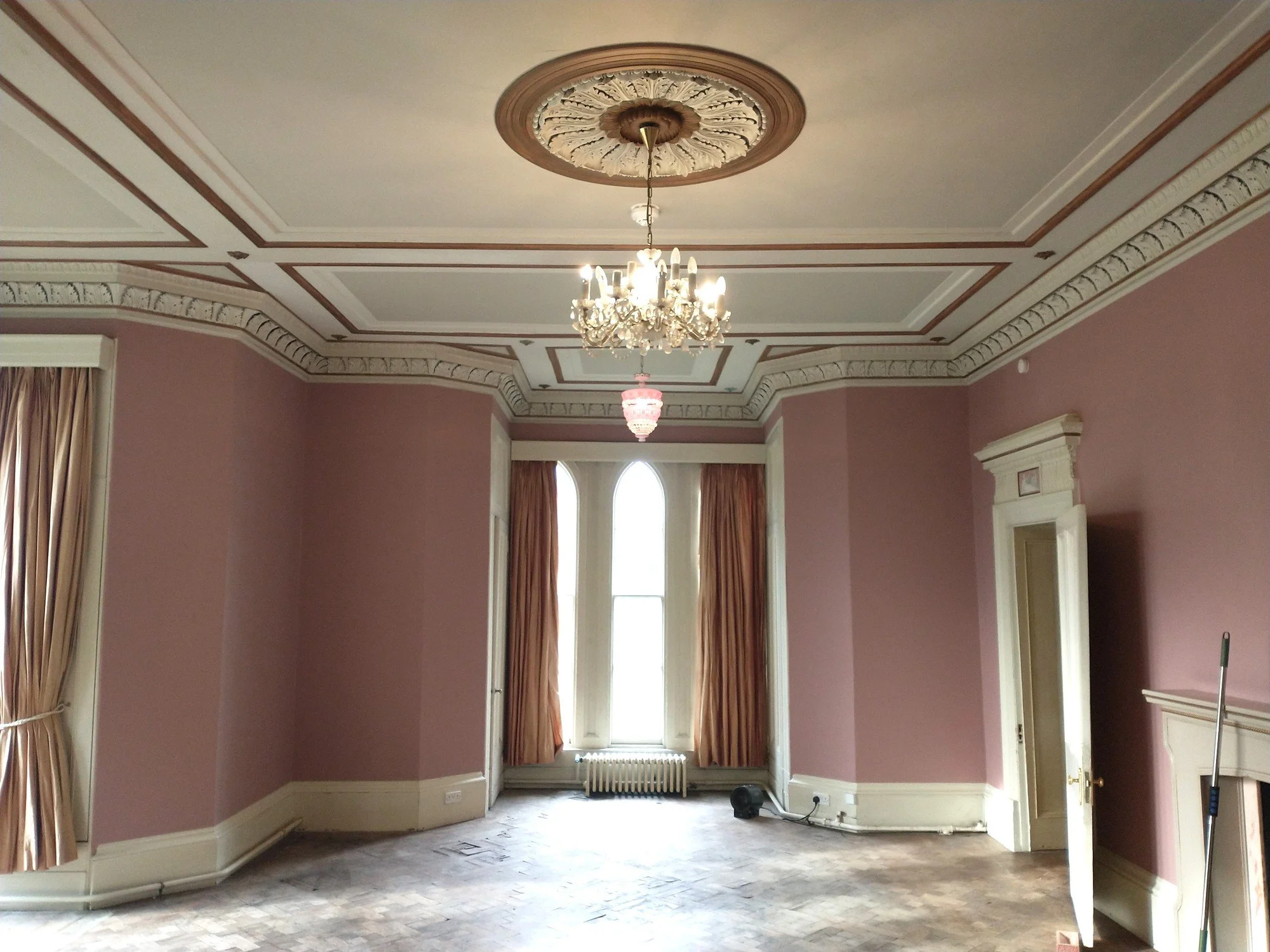 Redecorated reception room with soft pink walls, white woodwork, decorative cornicing and an ornate ceiling with chandeliers.