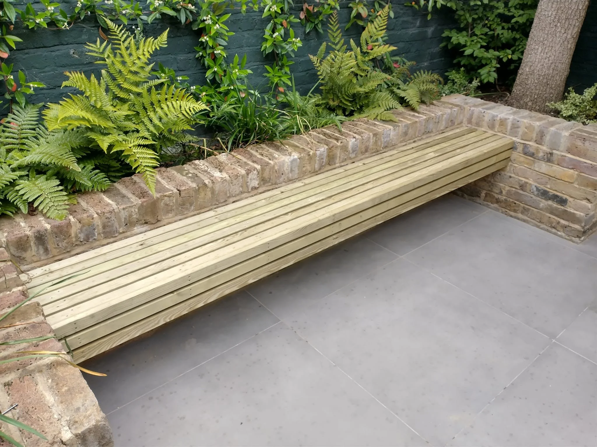 Slatted timber garden bench fitted into existing brickwork beside ferns and a paved patio.