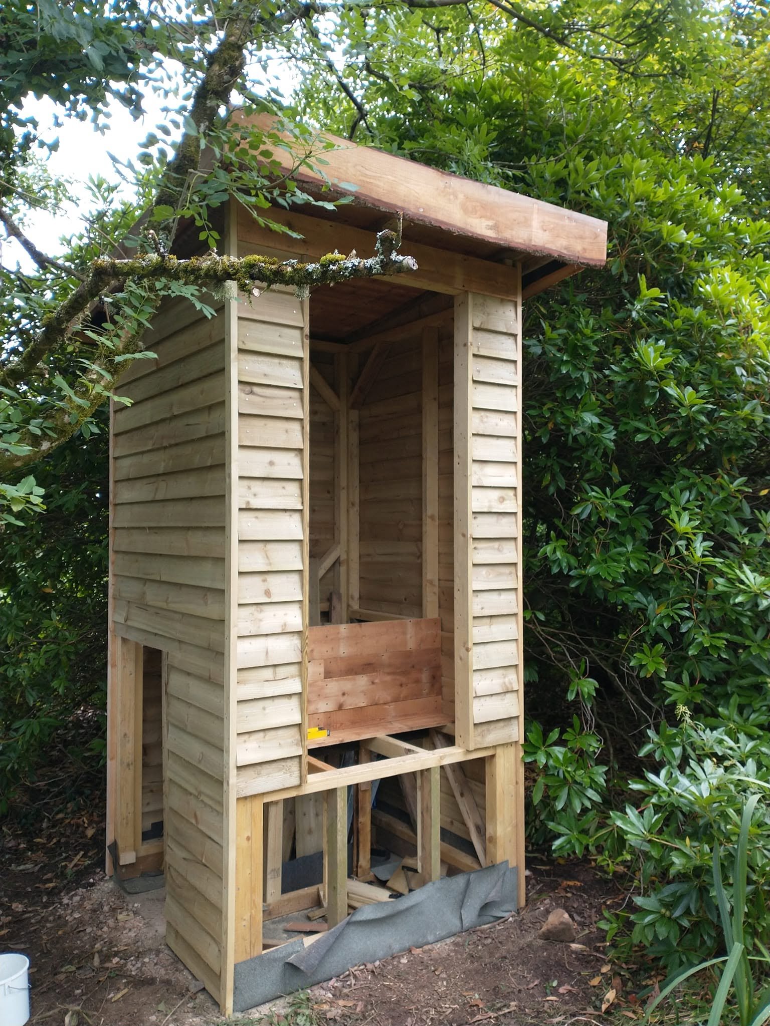 Composting toilet under construction, with timber cladding, a roof and an unfinished doorway.