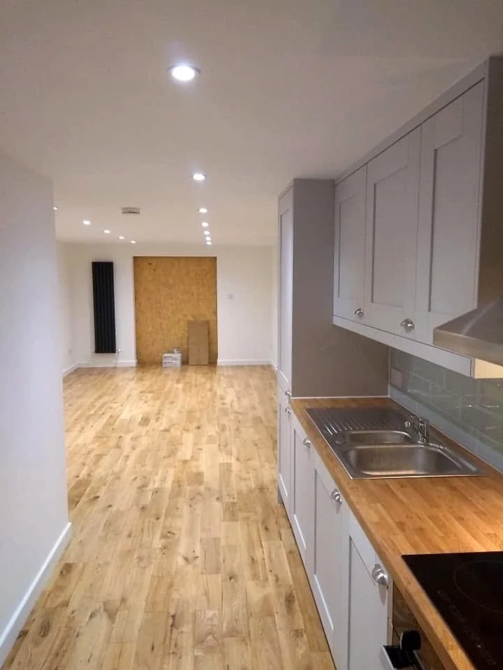 Converted garage interior with grey kitchen cabinets, timber-effect flooring, recessed ceiling lights and a boarded doorway.