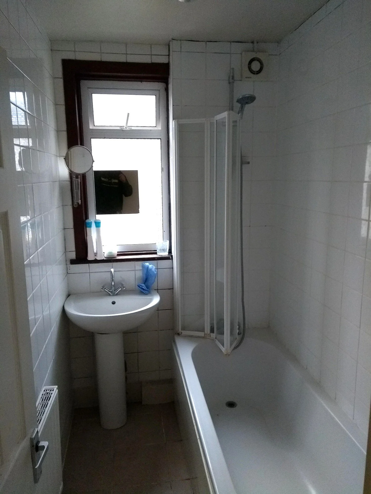 Narrow bathroom before refurbishment, with a pedestal basin, white tiled walls and a folding bath screen.