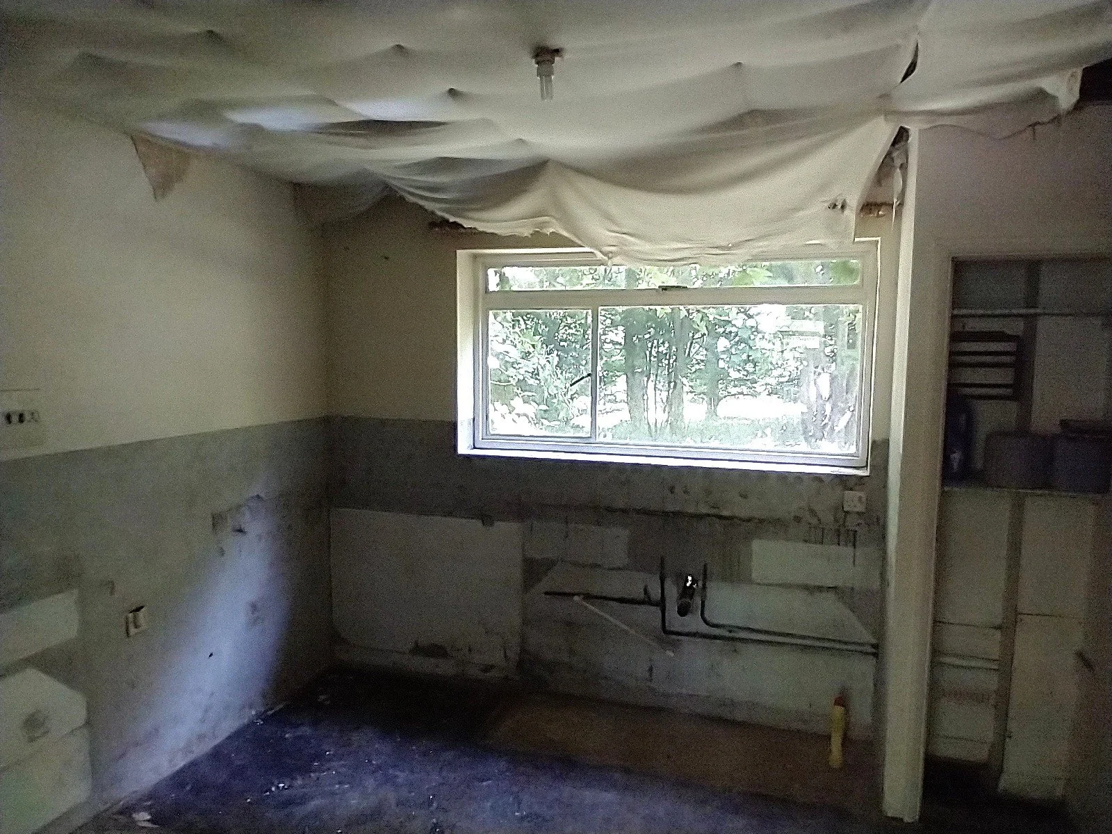 Stripped kitchen with exposed pipework, bare walls and loose ceiling covering before refurbishment.