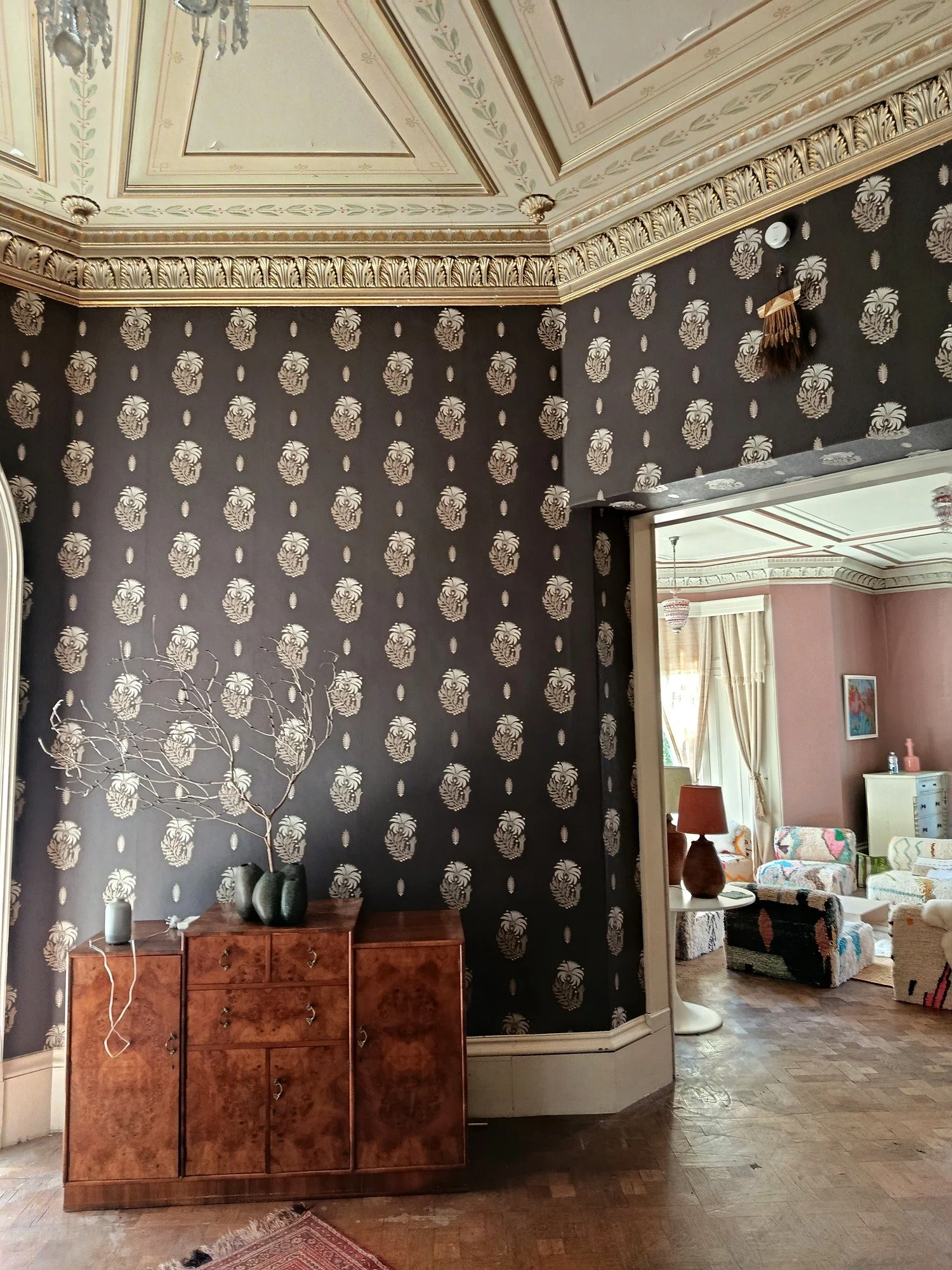 Patterned wallpaper around a wide doorway, with a wooden sideboard against the wall and a pink reception room beyond.