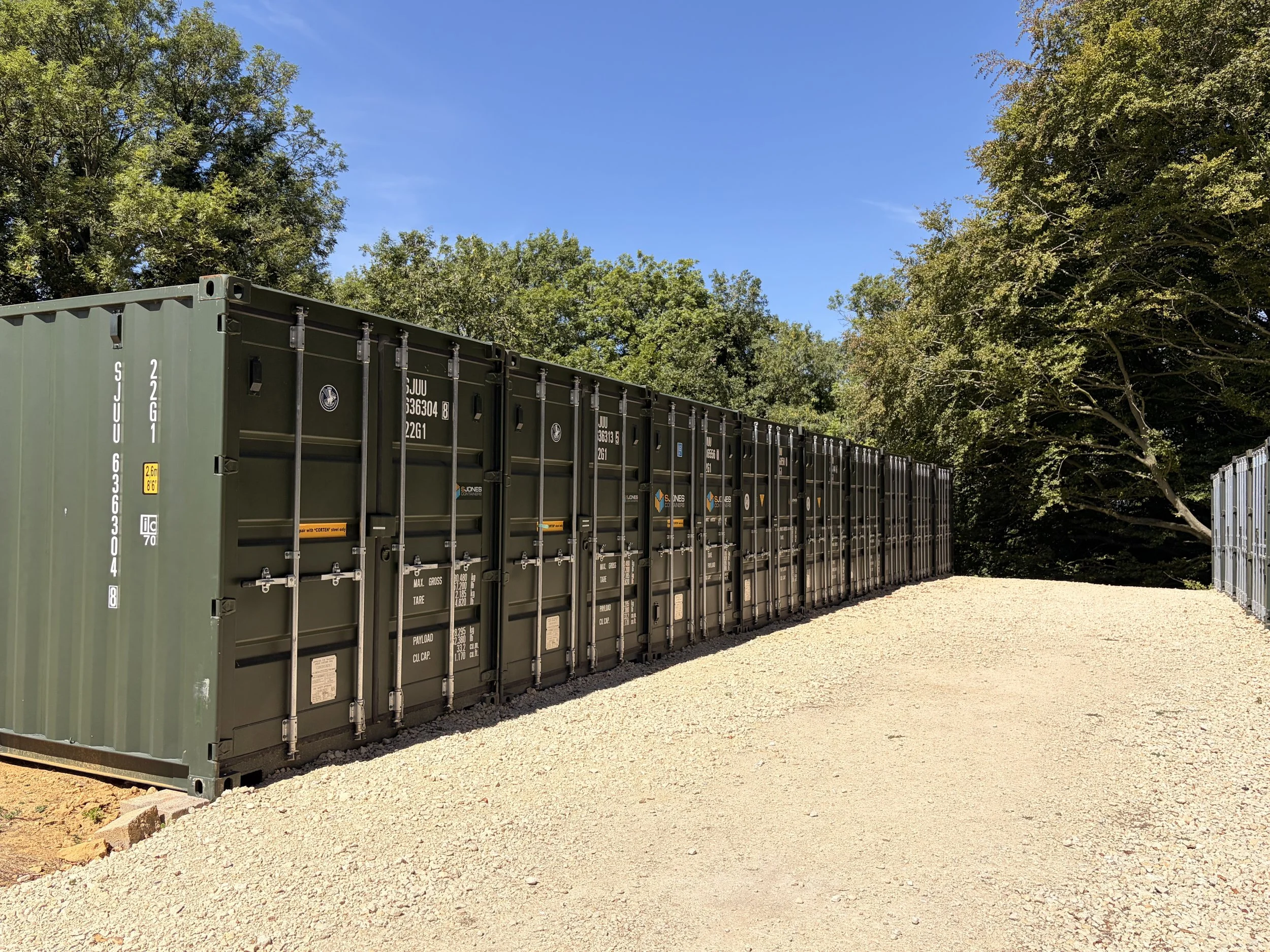 A row of green shipping containers lined up outdoors on a gravel ground, with trees and clear blue sky in the background.