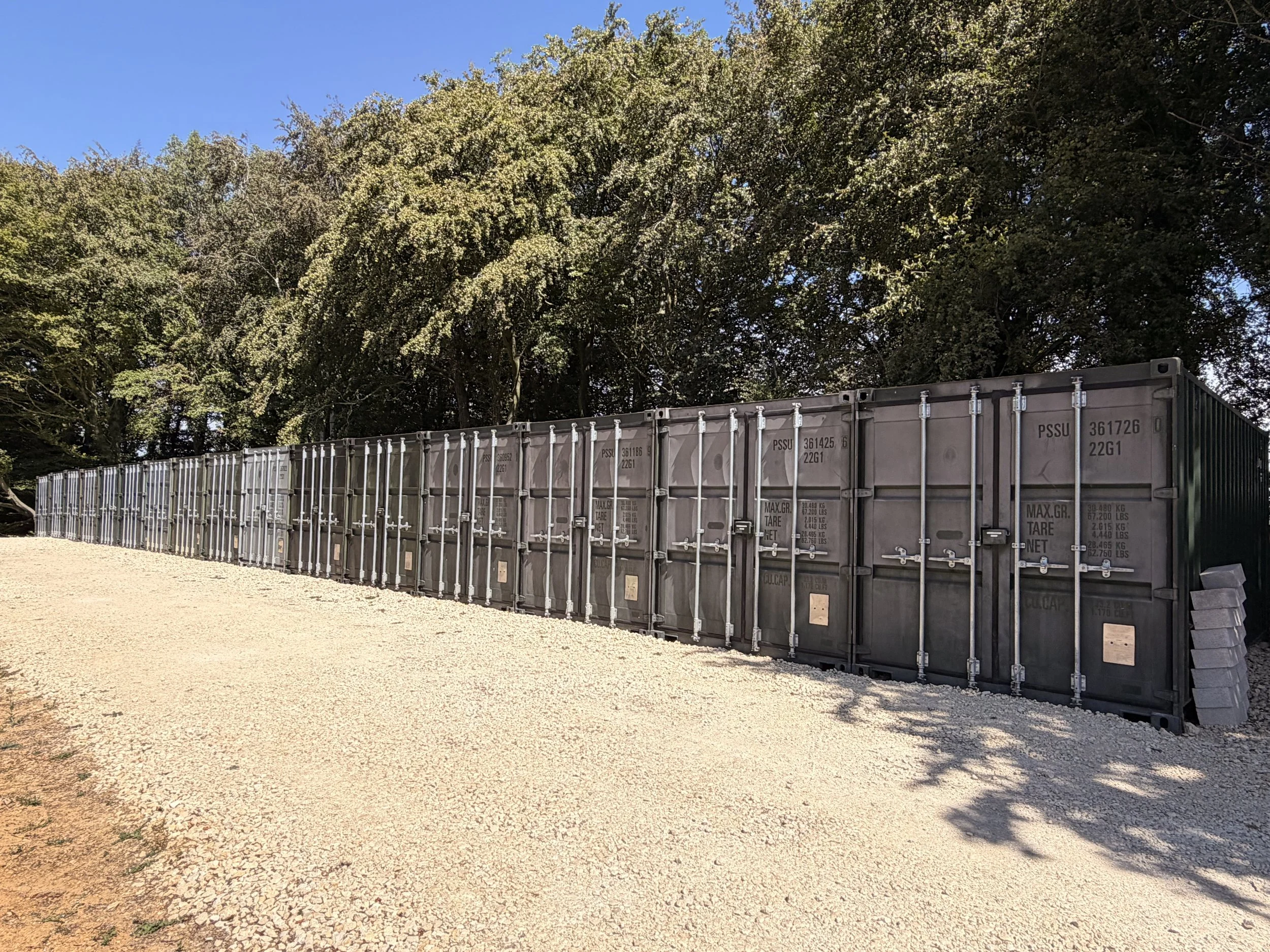 A row of gray shipping containers lined up on gravel ground, with a backdrop of dense trees and a clear blue sky.
