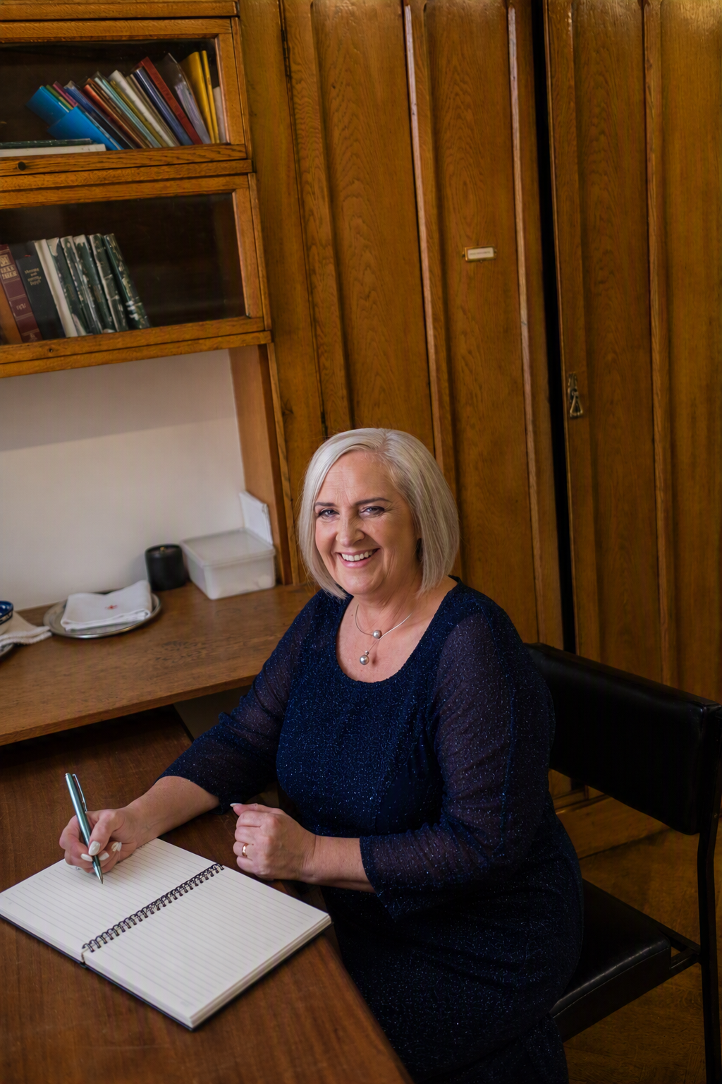 An older woman with blonde hair sitting at a wooden desk in a room with wooden cabinets and shelves. She is smiling, holding a pen, and writing in an open notebook.