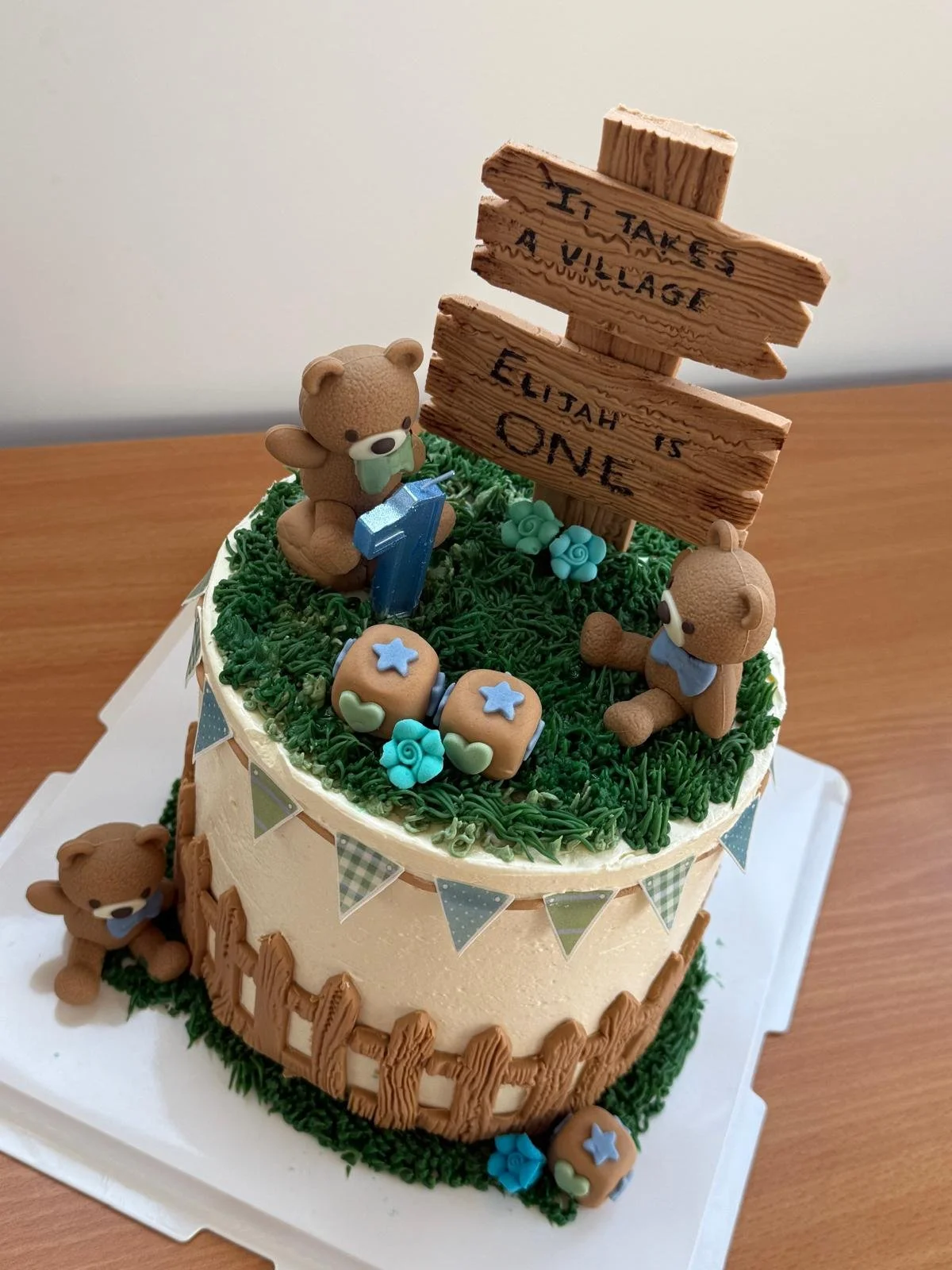 Teddy bear first birthday cake with a wooden It Takes A Village sign, bunting and picket fence