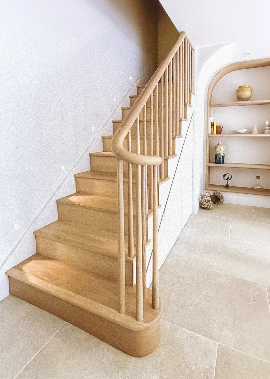 Bespoke oak staircase with a curved handrail and spindle balustrade, leading down to a limestone-floored basement with arched oak shelving, in Clifton by JG Build.