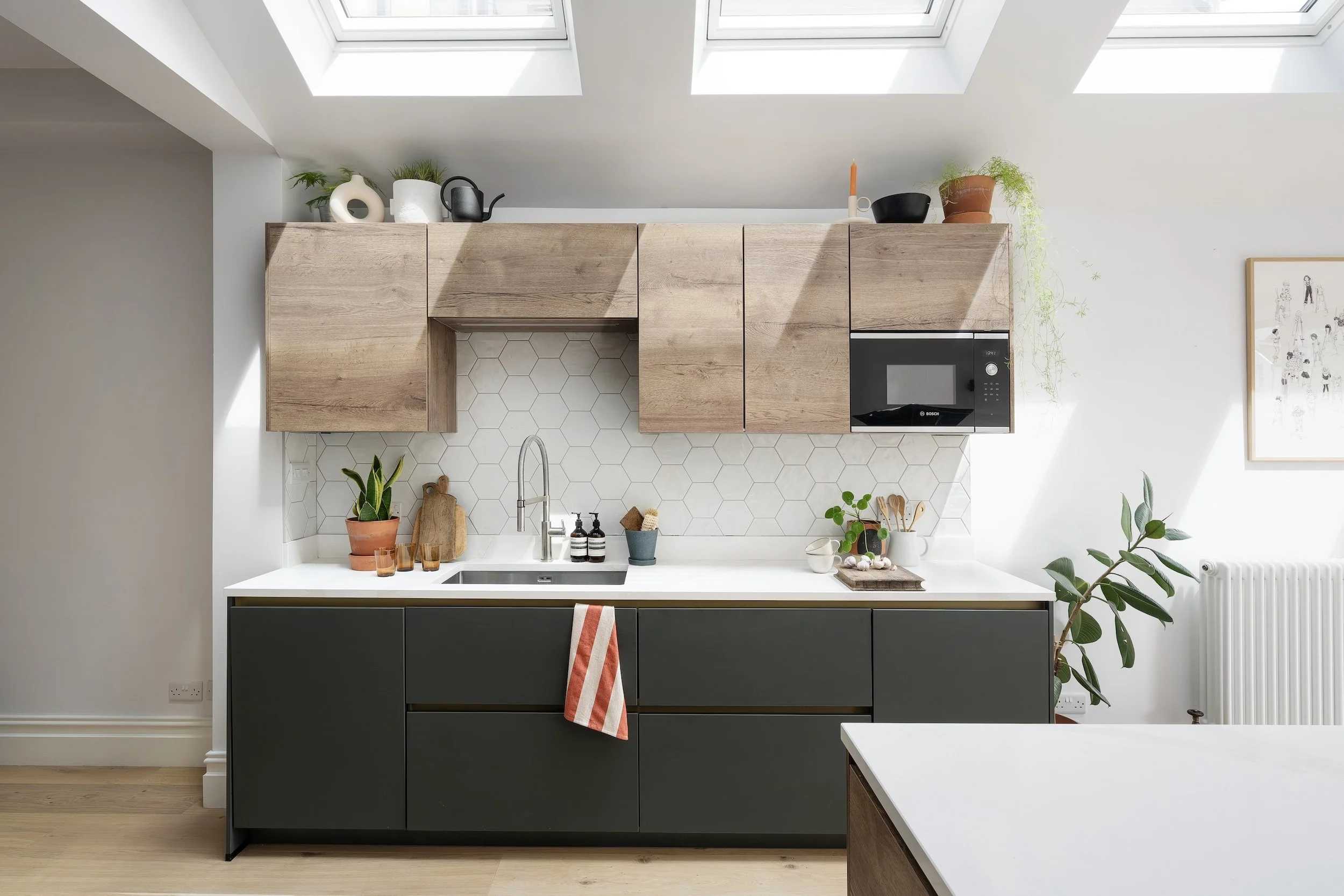 Green handleless base units and oak-effect wall cabinets with a white hexagonal tiled splashback, beneath a row of roof lights, in North Bristol by JG Build.