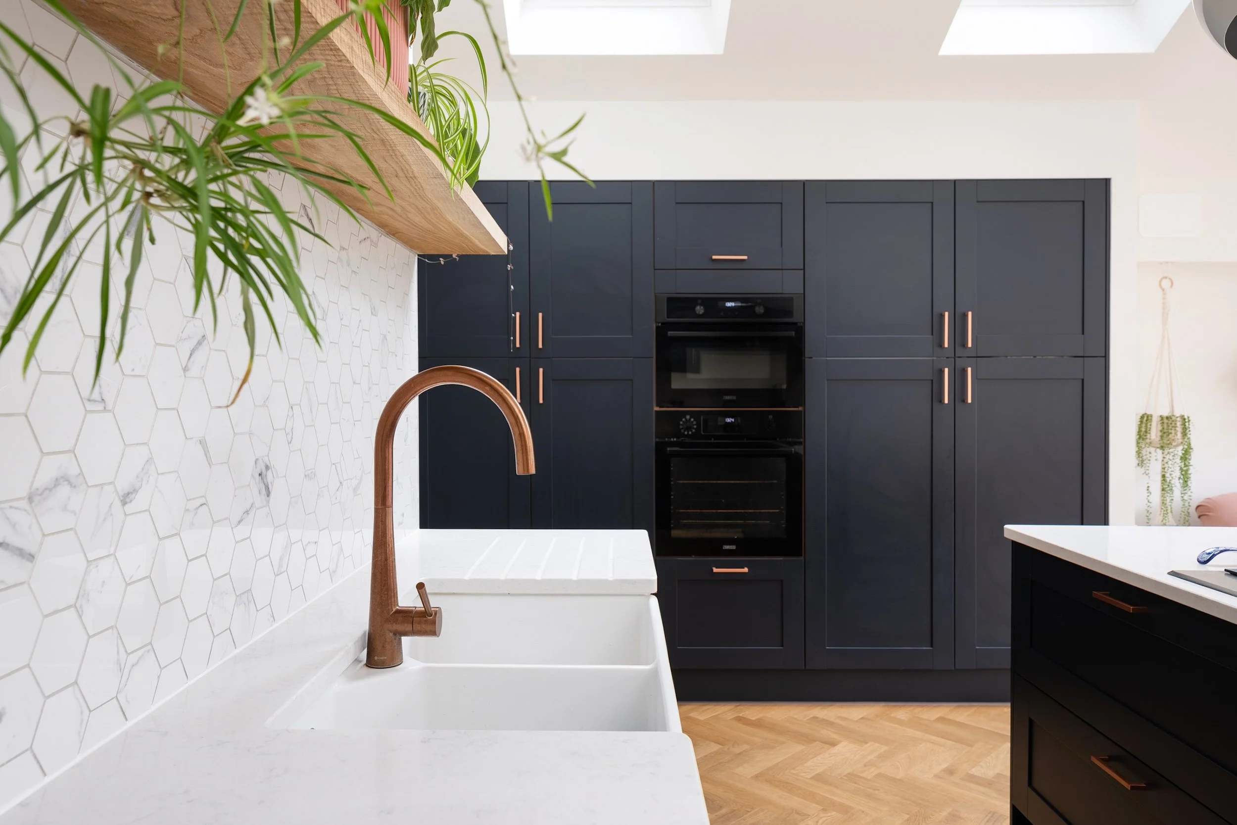 Copper tap and white ceramic sink against a marble-effect hexagonal splashback, with tall navy shaker units and built-in ovens behind, in Bishopston by JG Build.