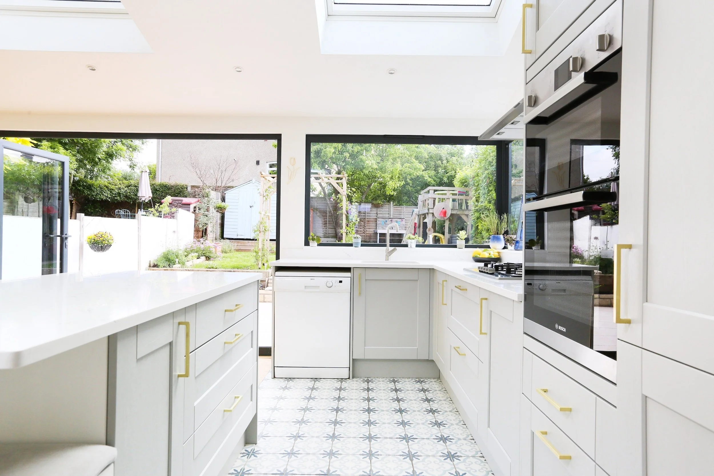 Pale grey shaker kitchen with gold handles and patterned floor tiles, with a large picture window and bi-fold doors looking onto the garden, in Bristol by JG Build.